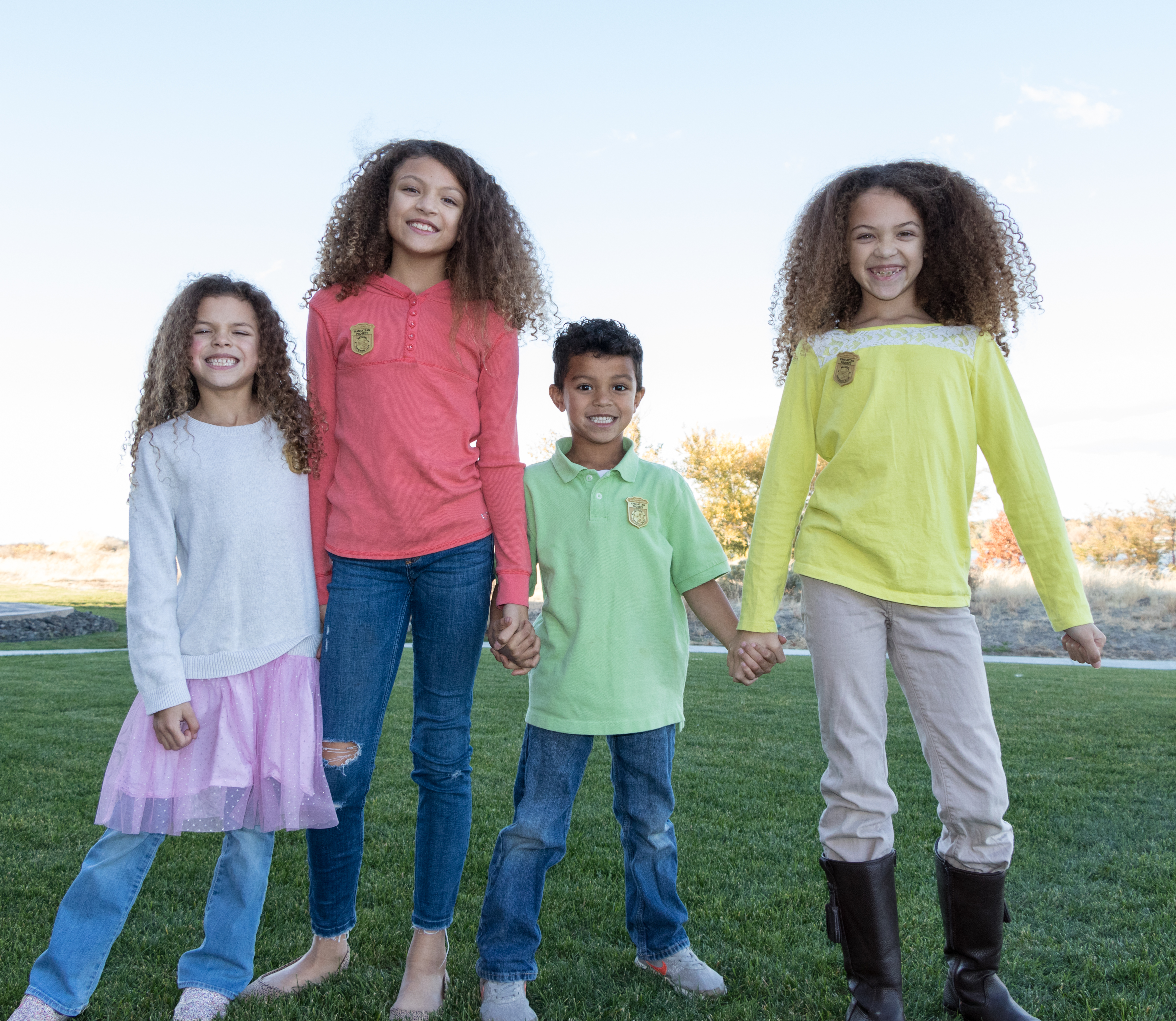 three girls and a boy smiling and holding hands stand on green grass.
