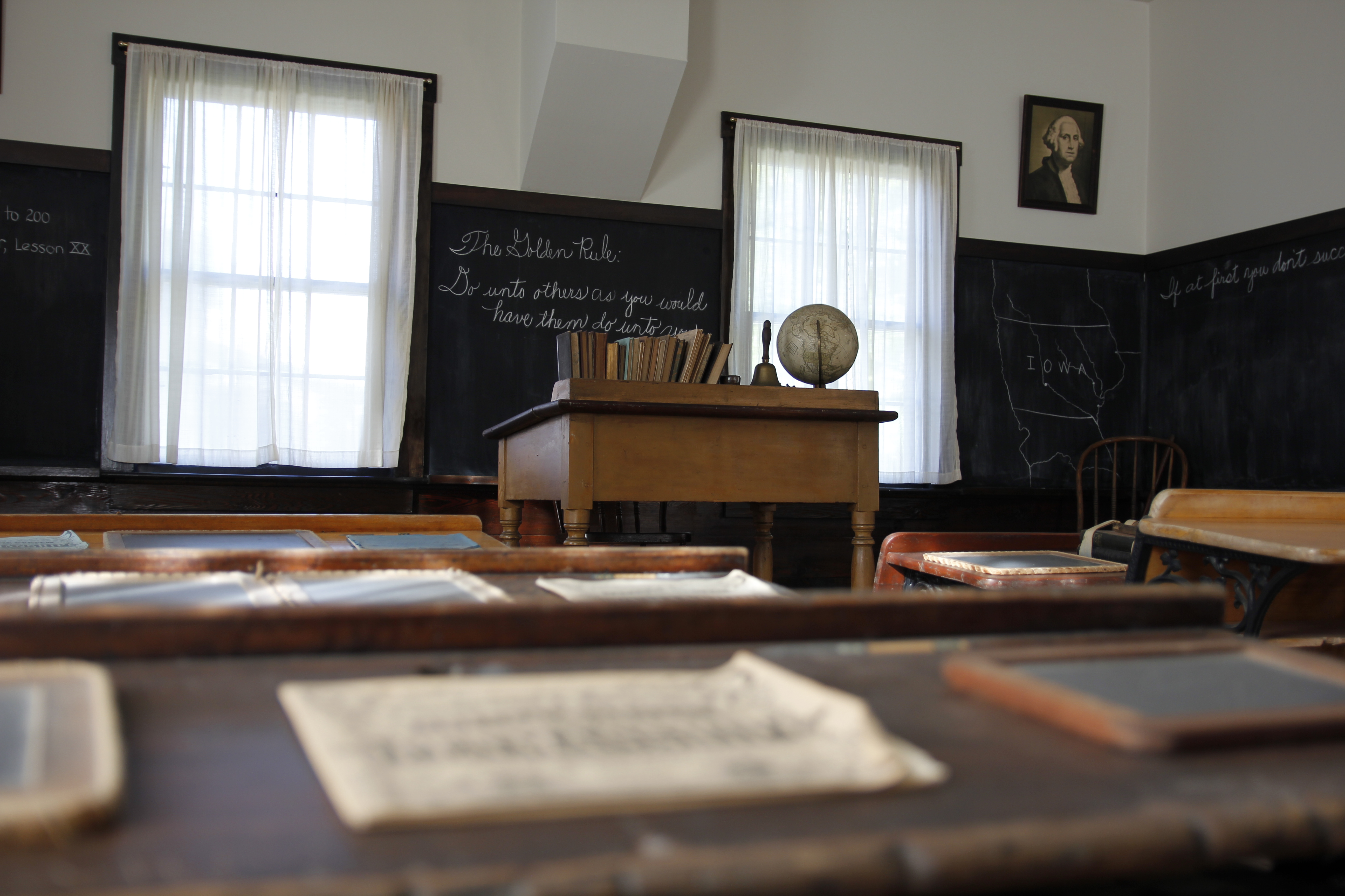 An 1870s classroom with an elevated teacher's desk and chalkboards as seen from a pupil's desk.