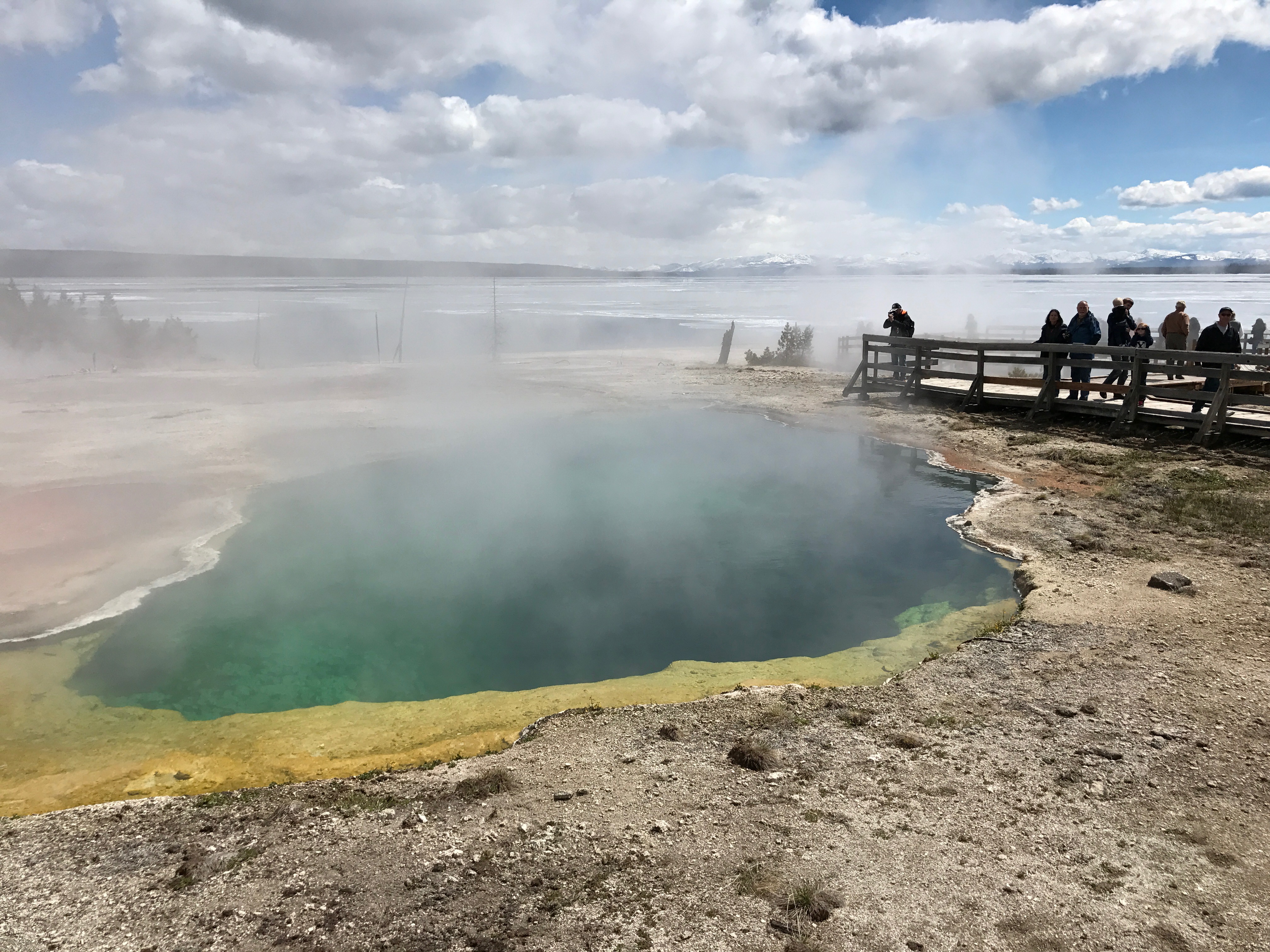 People stand on a boardwalk and photograph a steaming pool of water. A large large looms behind.