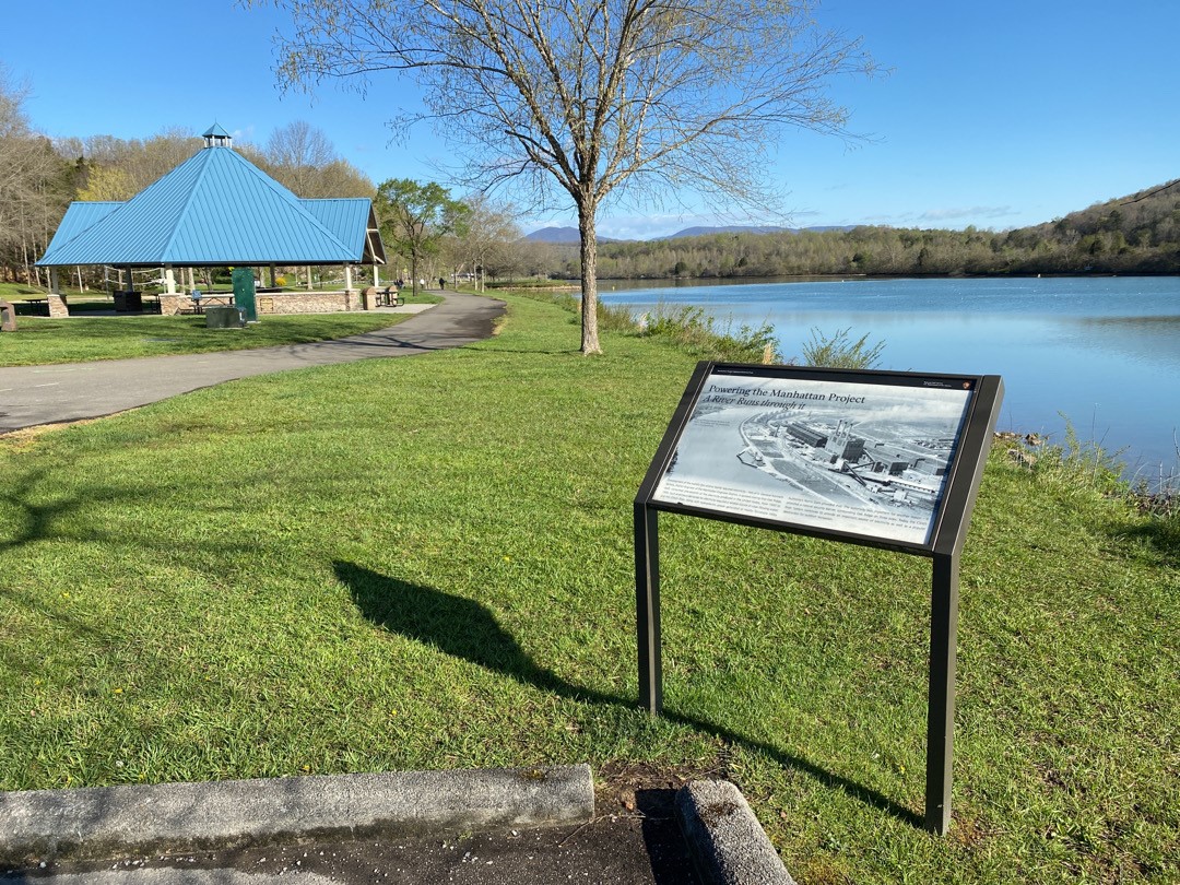A wayside panel next to a river with a park rain shelter in the background.