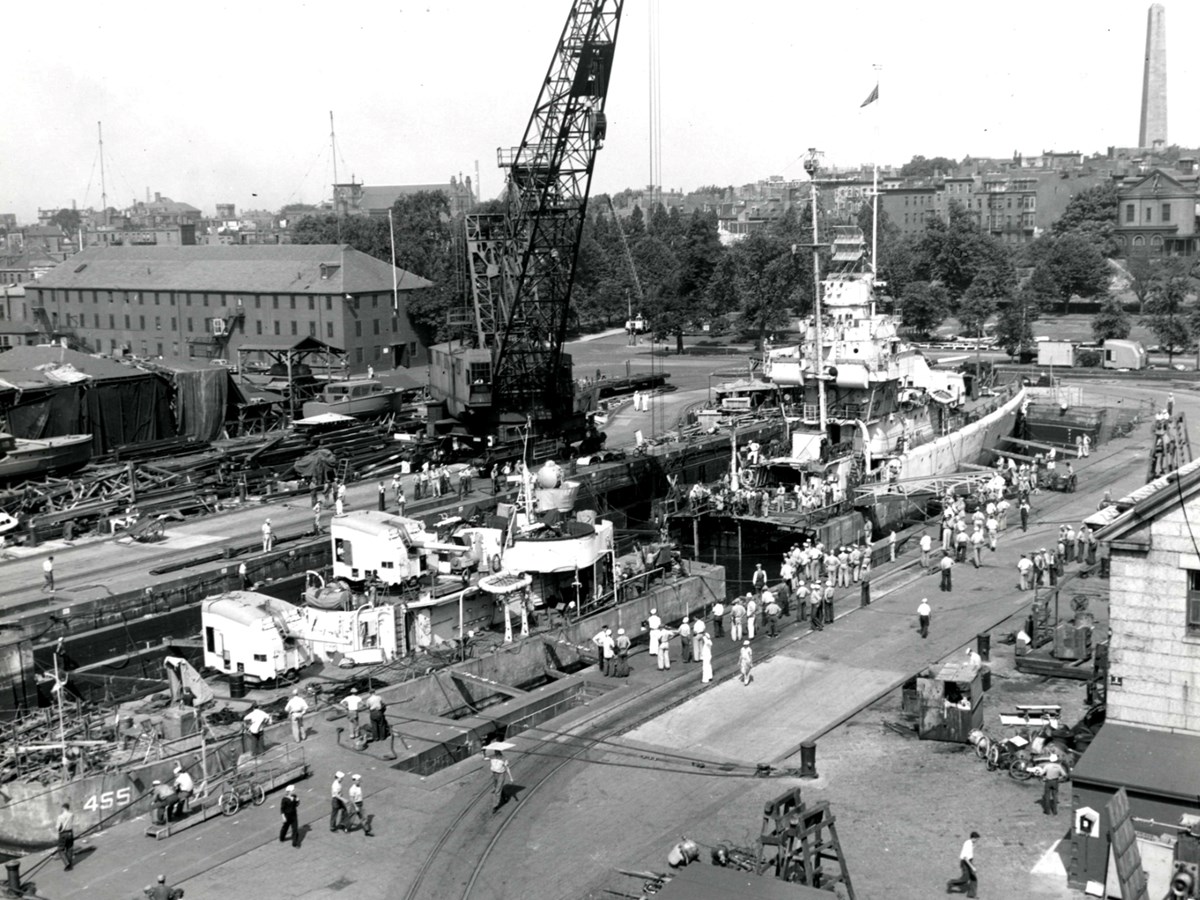black and white photo of a crane and a dry dock with a ship in it. Workers are seen throughout.