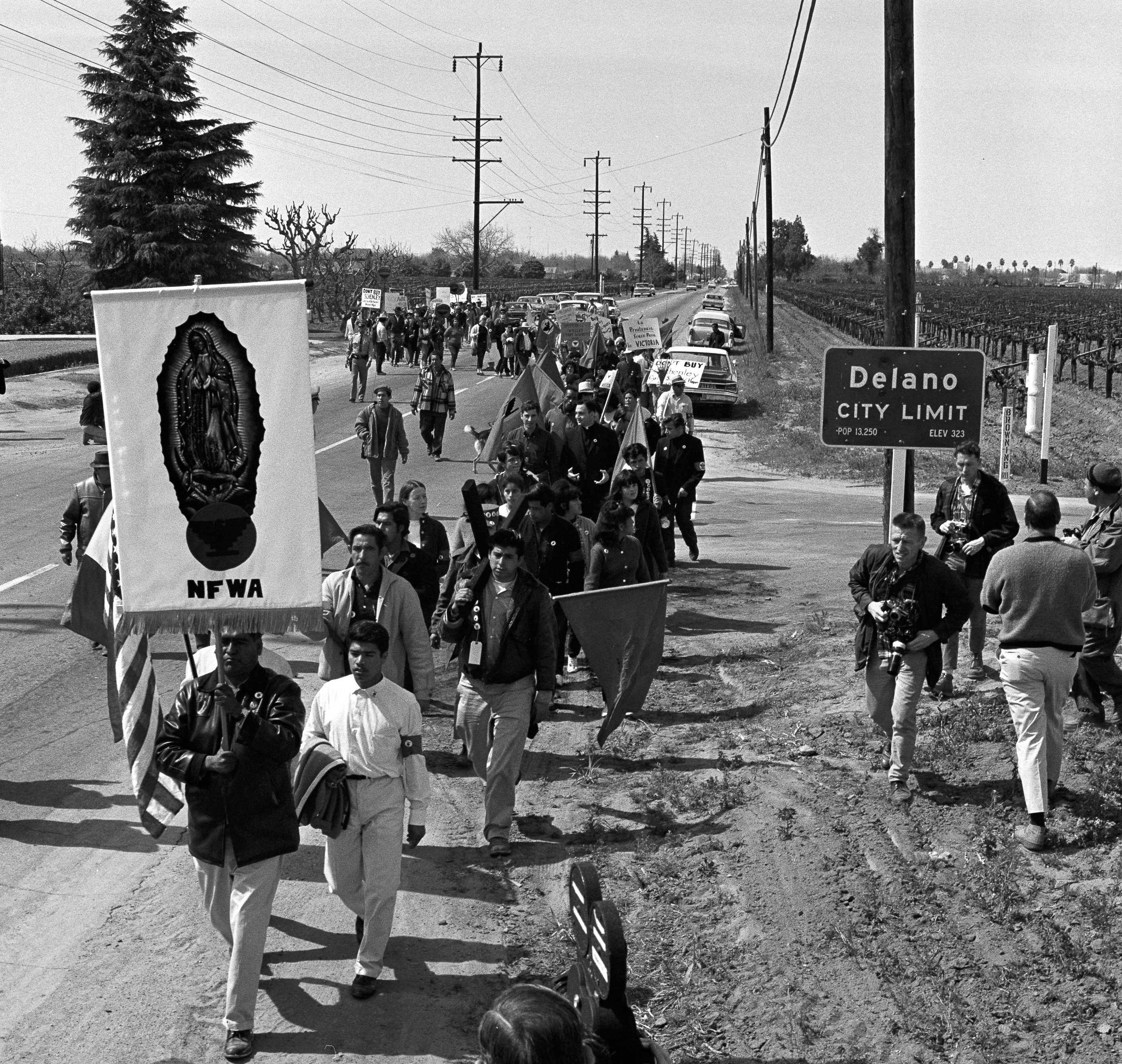 A long line of people walk along a road past a sign for the city limits of Delano, California. Some