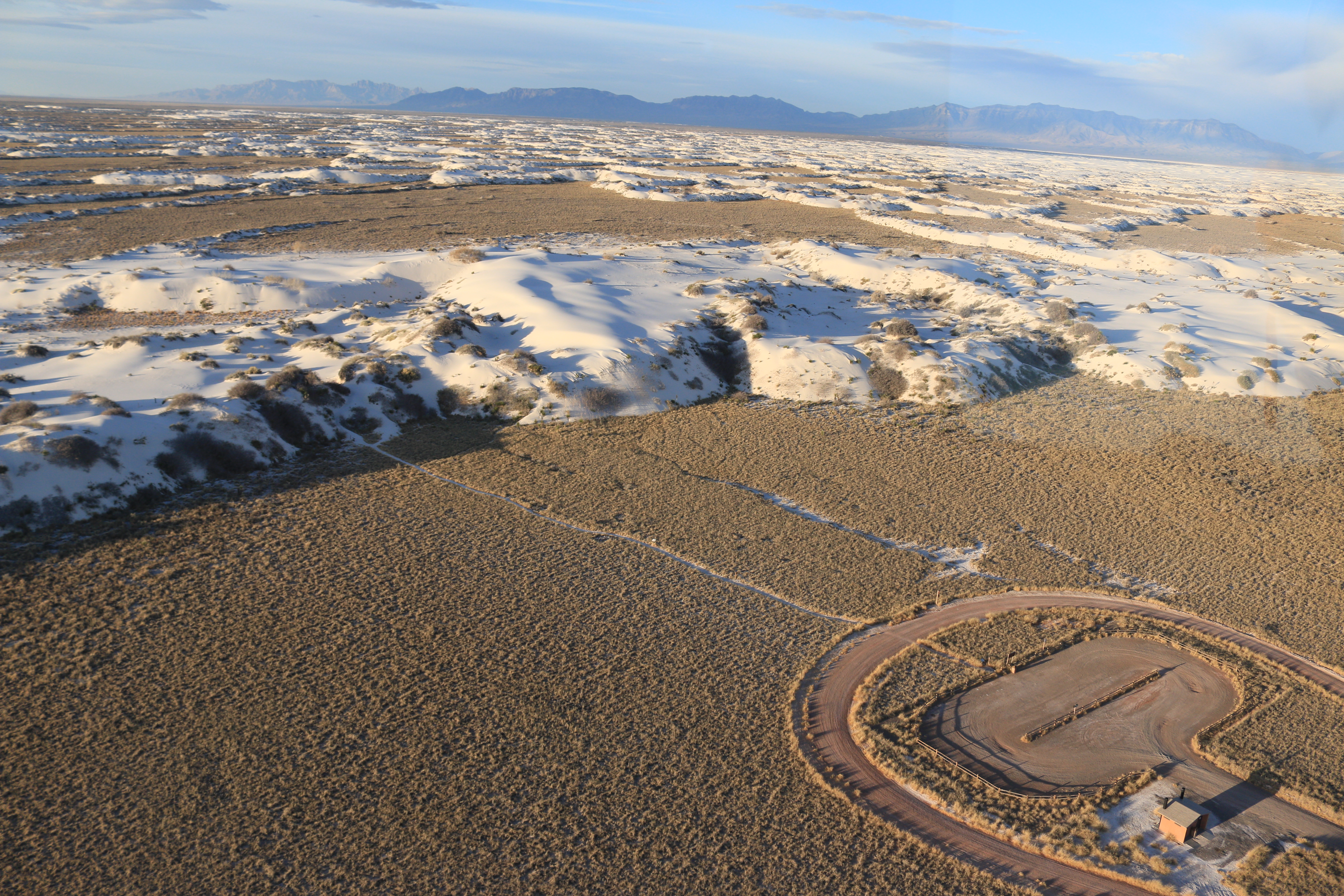 Aerial photo of white dunes meeting brown desert with a parking lot in the bottom right corner.