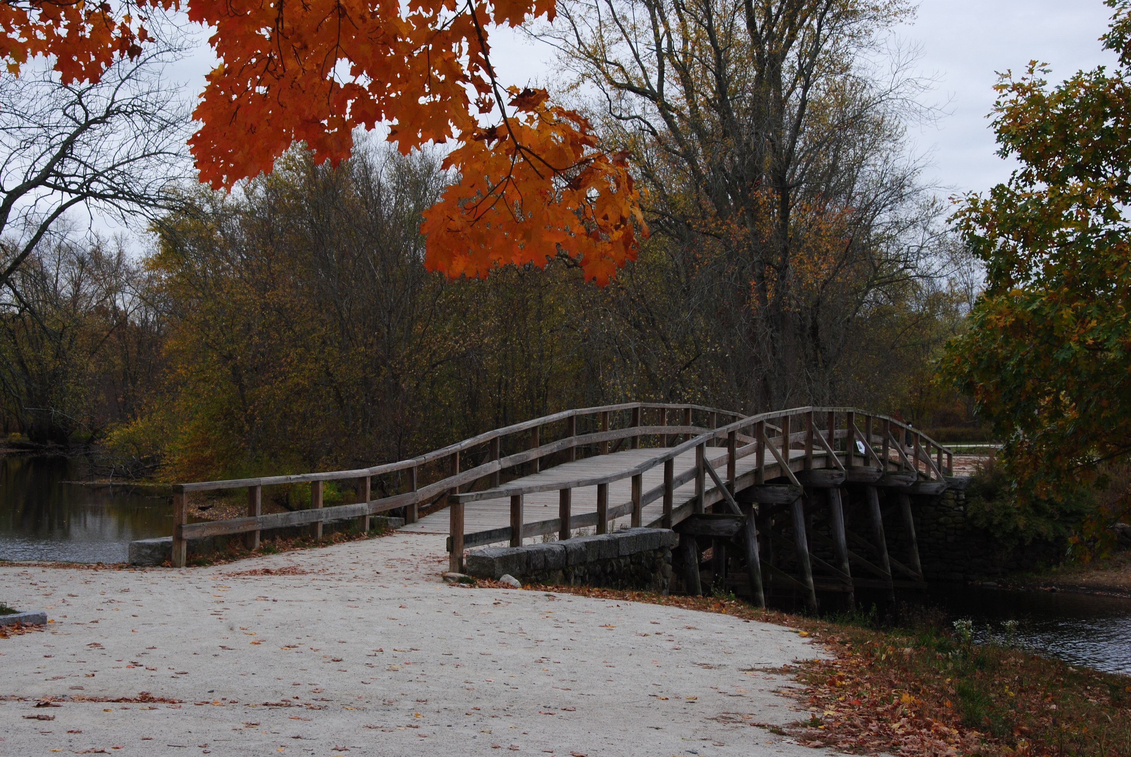 An arched wooden bridge spans a small river. Trees display autumn color against an overcast sky.