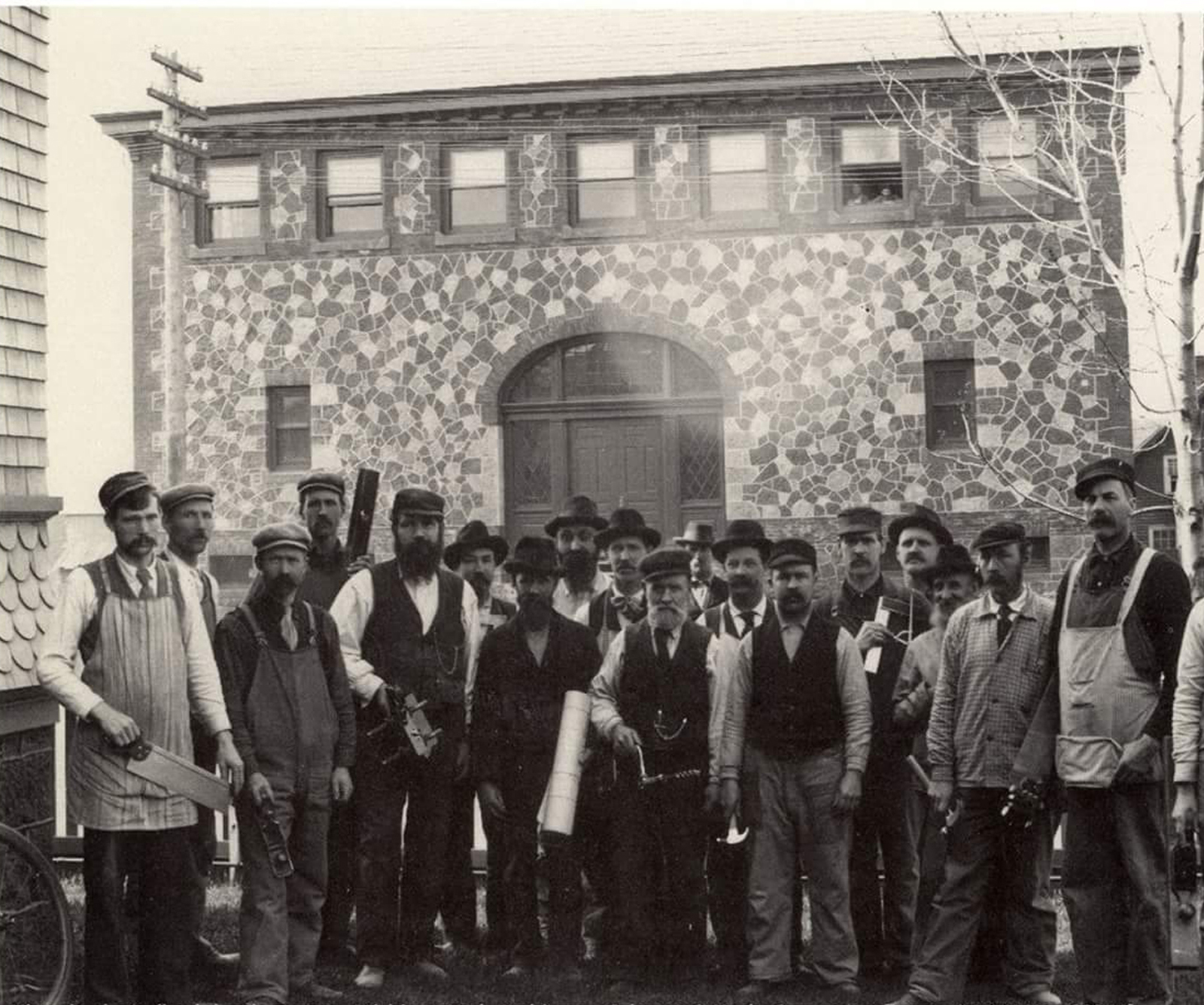 Black and white image with men posing with tools in front of a stone two story building.