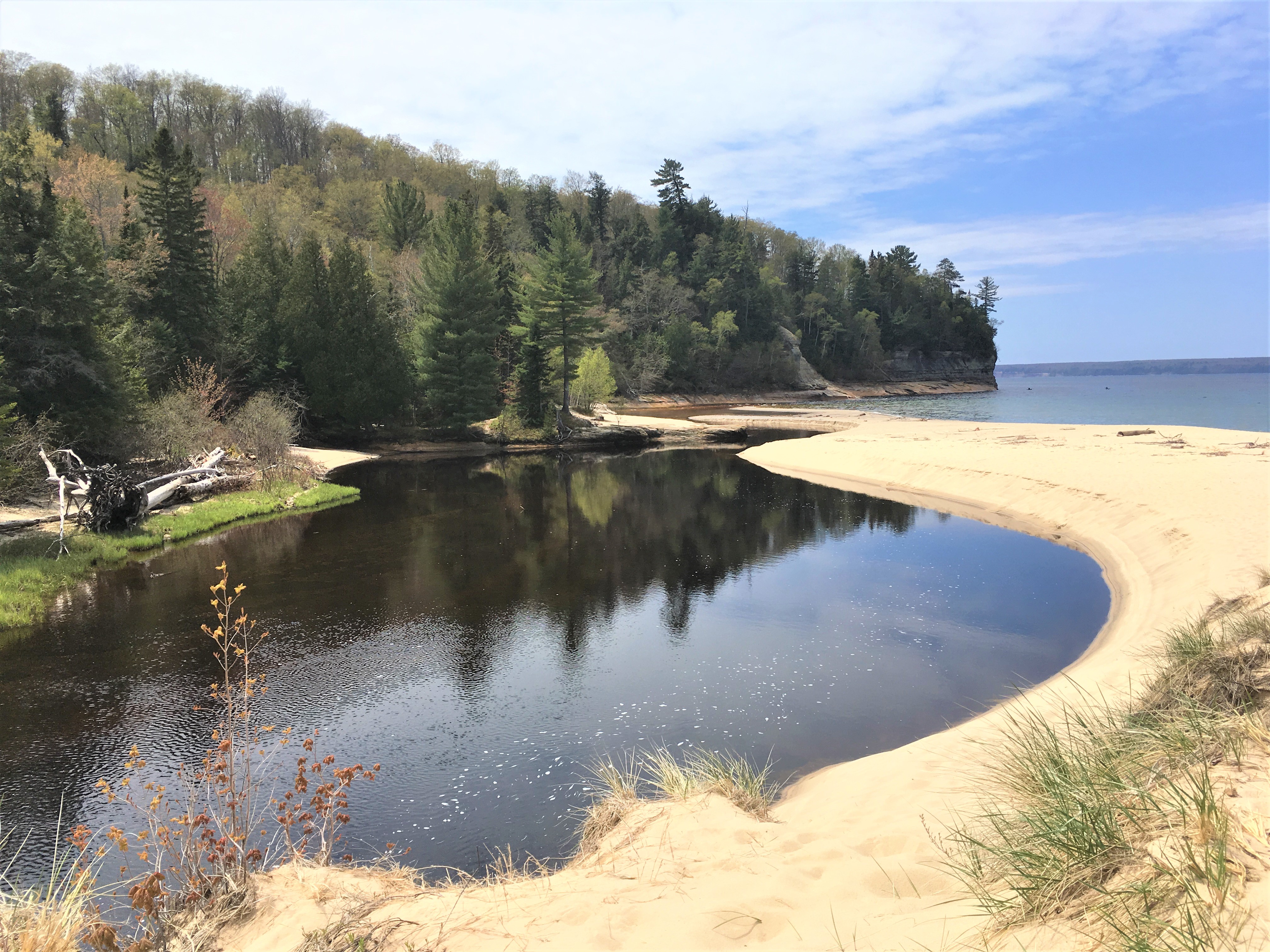 River flowing across sand beach into Lake Superior with forest cliff slope in the background