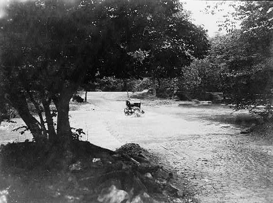 A black and white image of a car driving through a ford in a creek