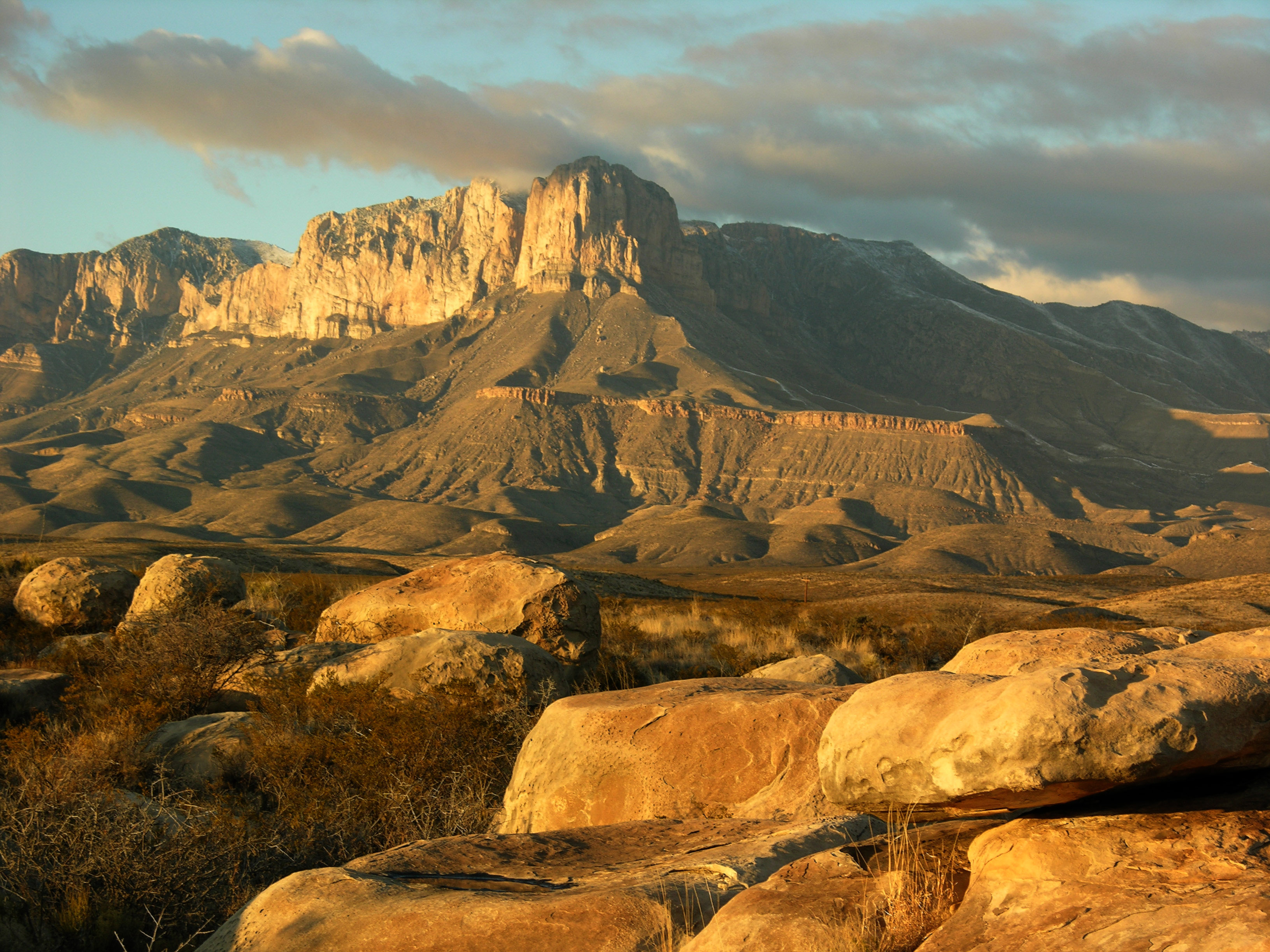 Orange hued mountain range above rolling hills under a cloudy blue sky.