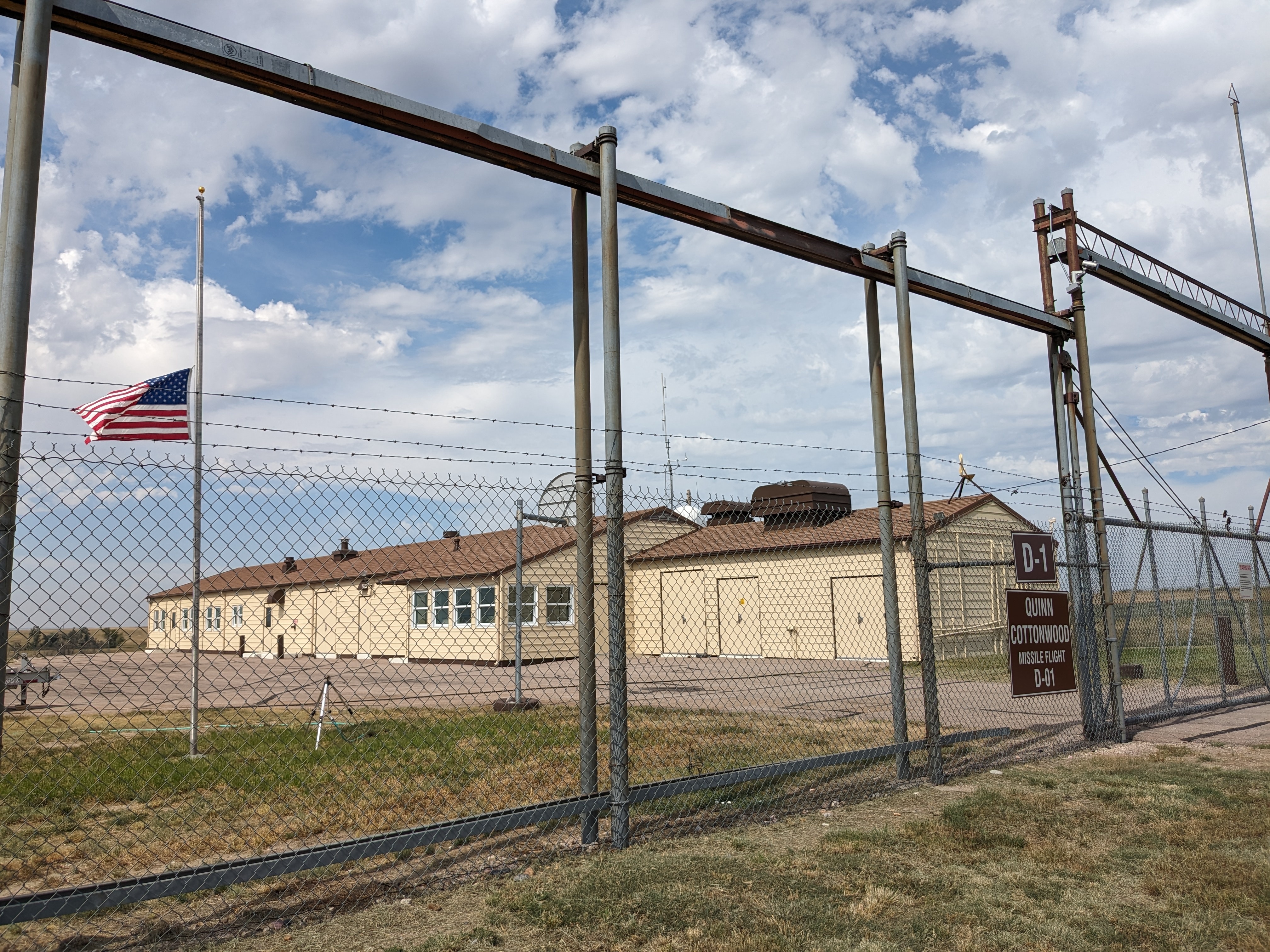 Long beige building behind a chain link fence. The flag on the pole in front of it is flying at half