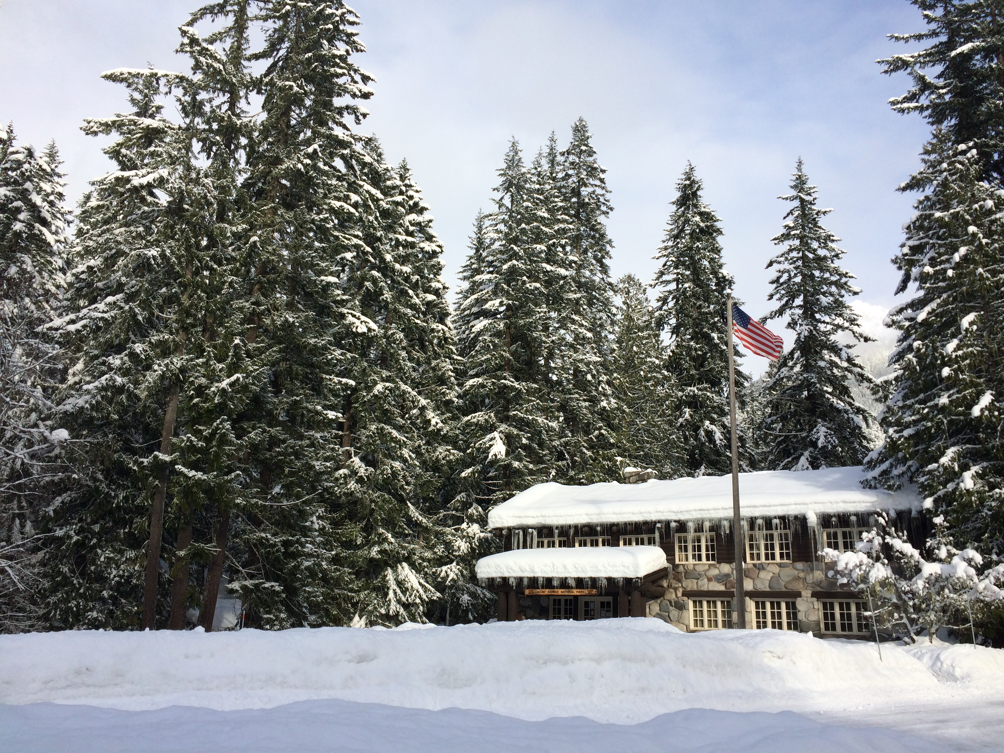 A two-story wood and stone building under a blanket of snow and surrounded by snowy forest.