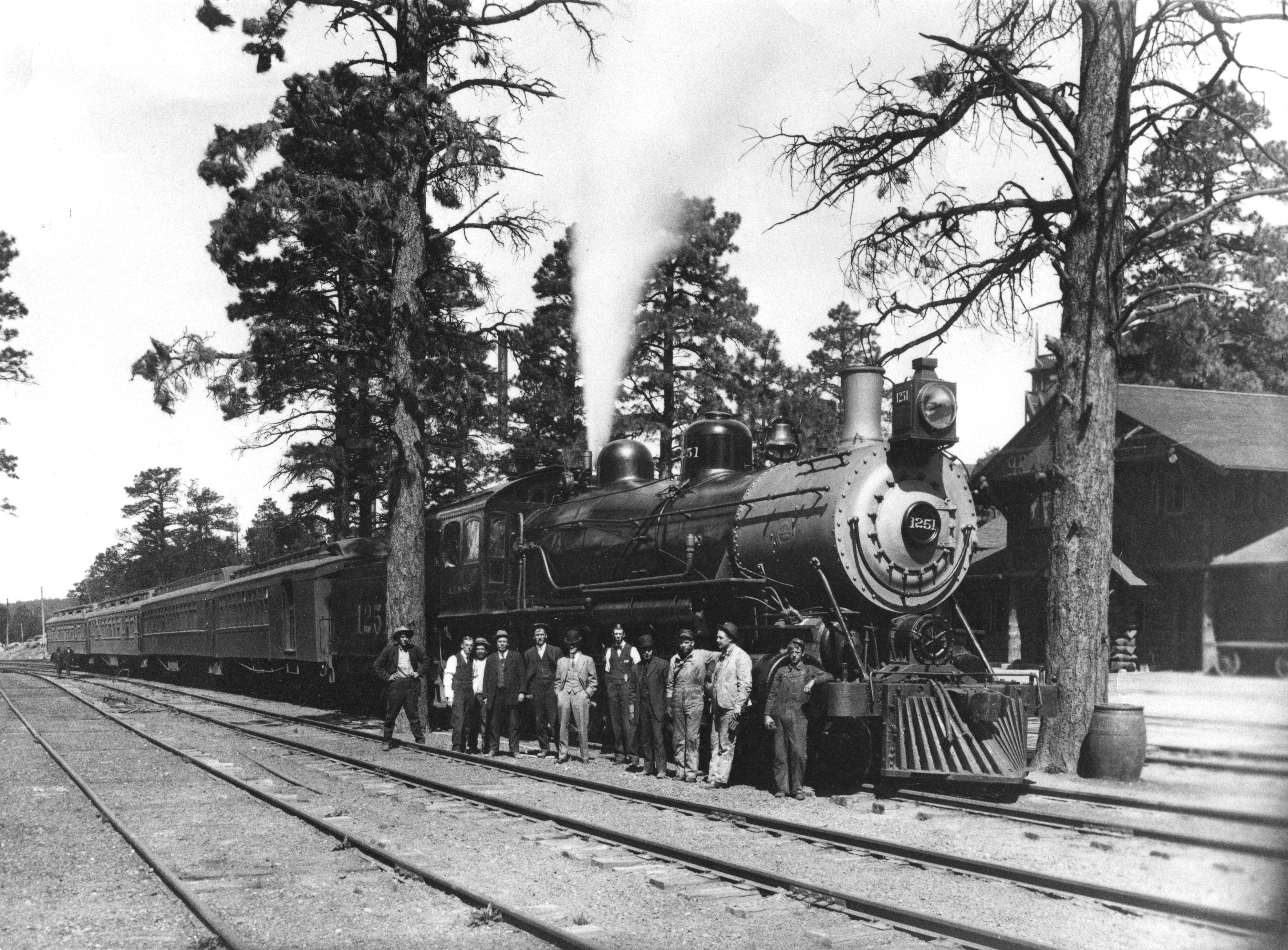 Black and white photo of 11 men standing in a line outside of a steam engine carrying 4 train cars.
