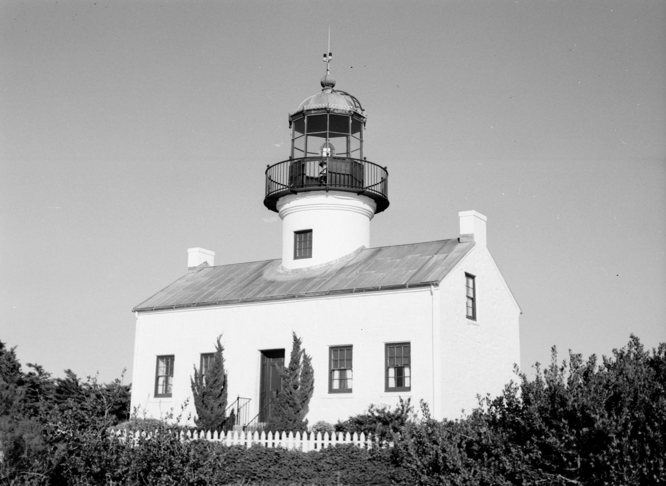 A black and white photo. A two-story building with a lighthouse tower sits nestles in bushes.