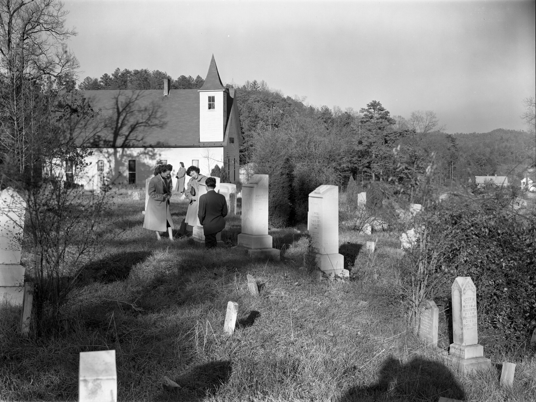 A black and white photograph of a small church and graveyard on a wooded hillside.