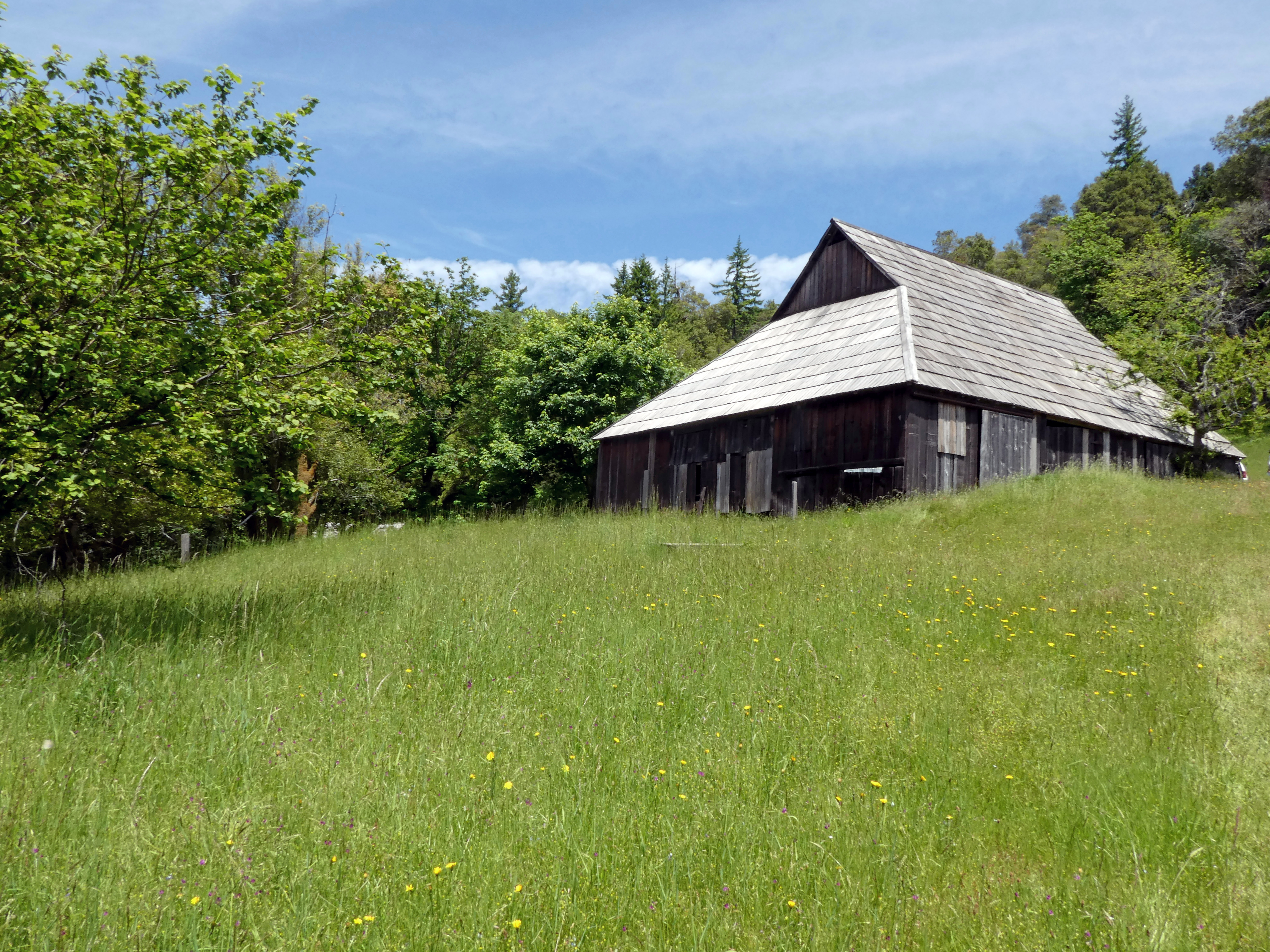 A grassy slope leads upto a wooden barn with a pitched roof. Trees surround the barn.