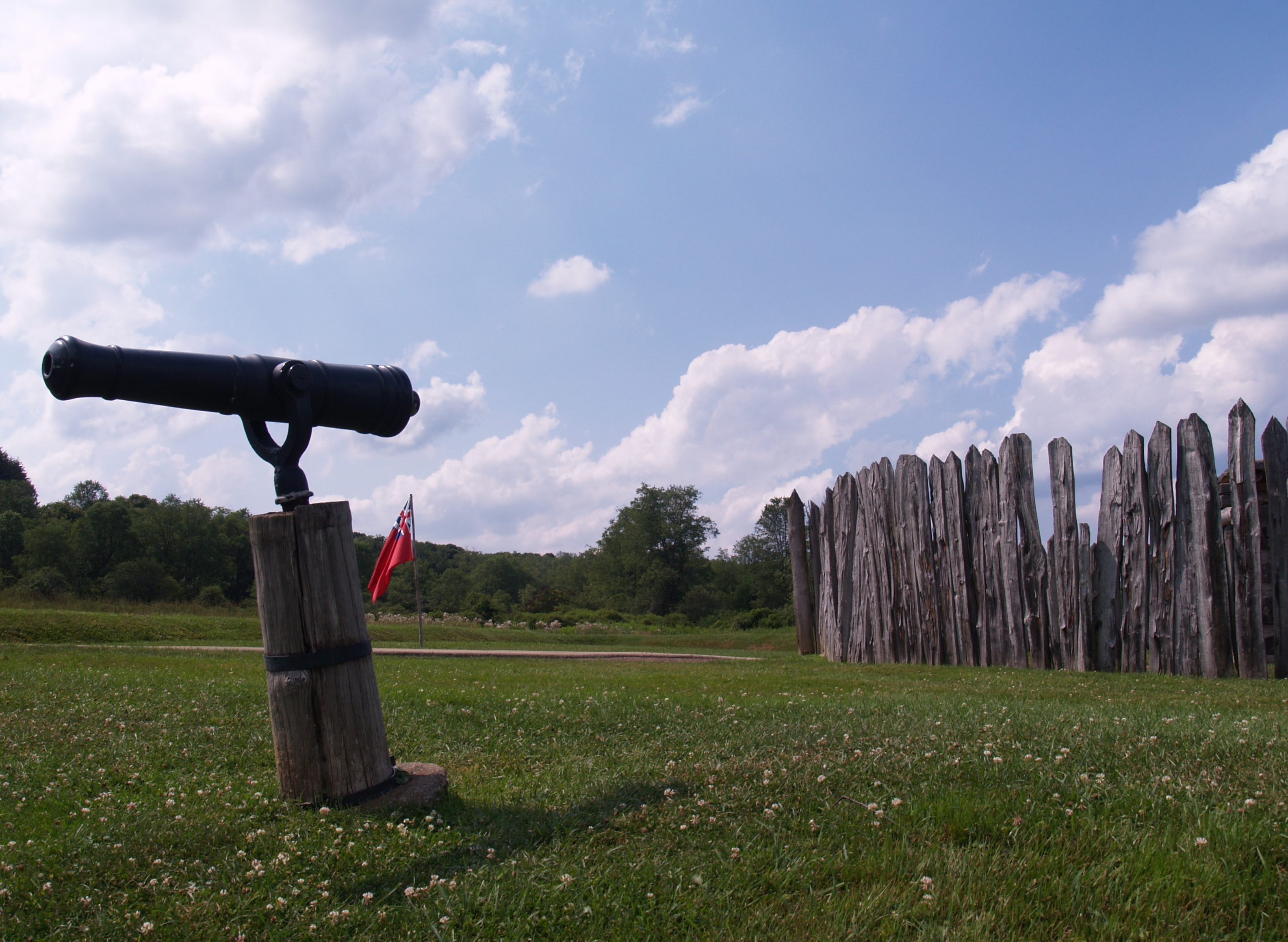 A swivel gun (small canon) stand in front of a small circular stockade fort with a red British flag.