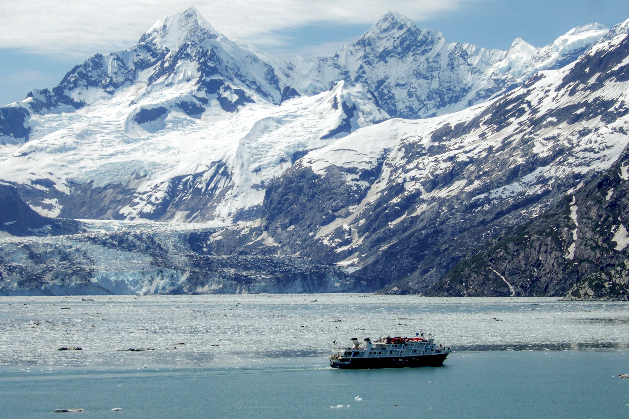 A tour boat is dwarfed by steep rugged icy mountains, while a glacier pours into water, overcast day