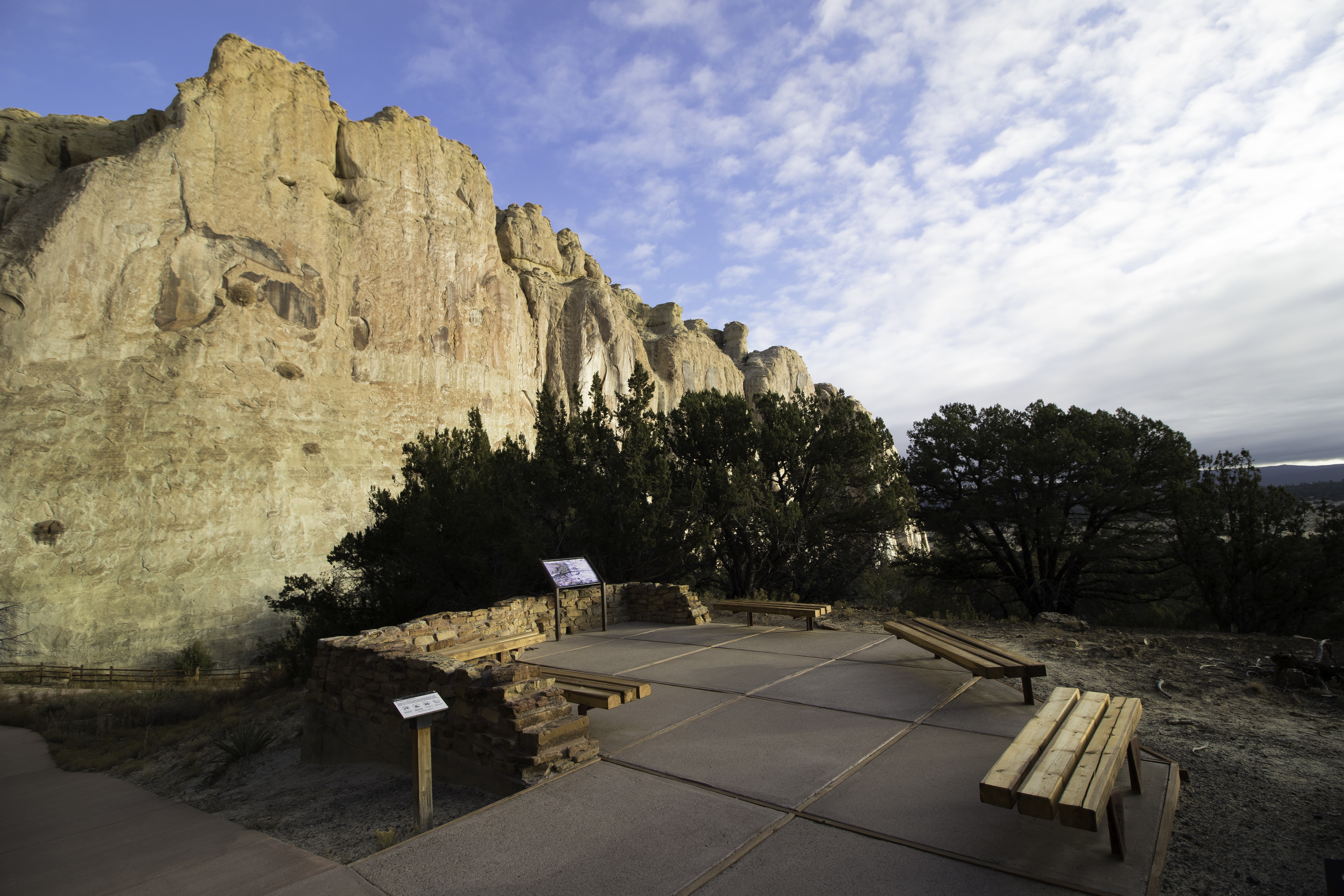 A patio with benches and juniper trees by a large cliff face