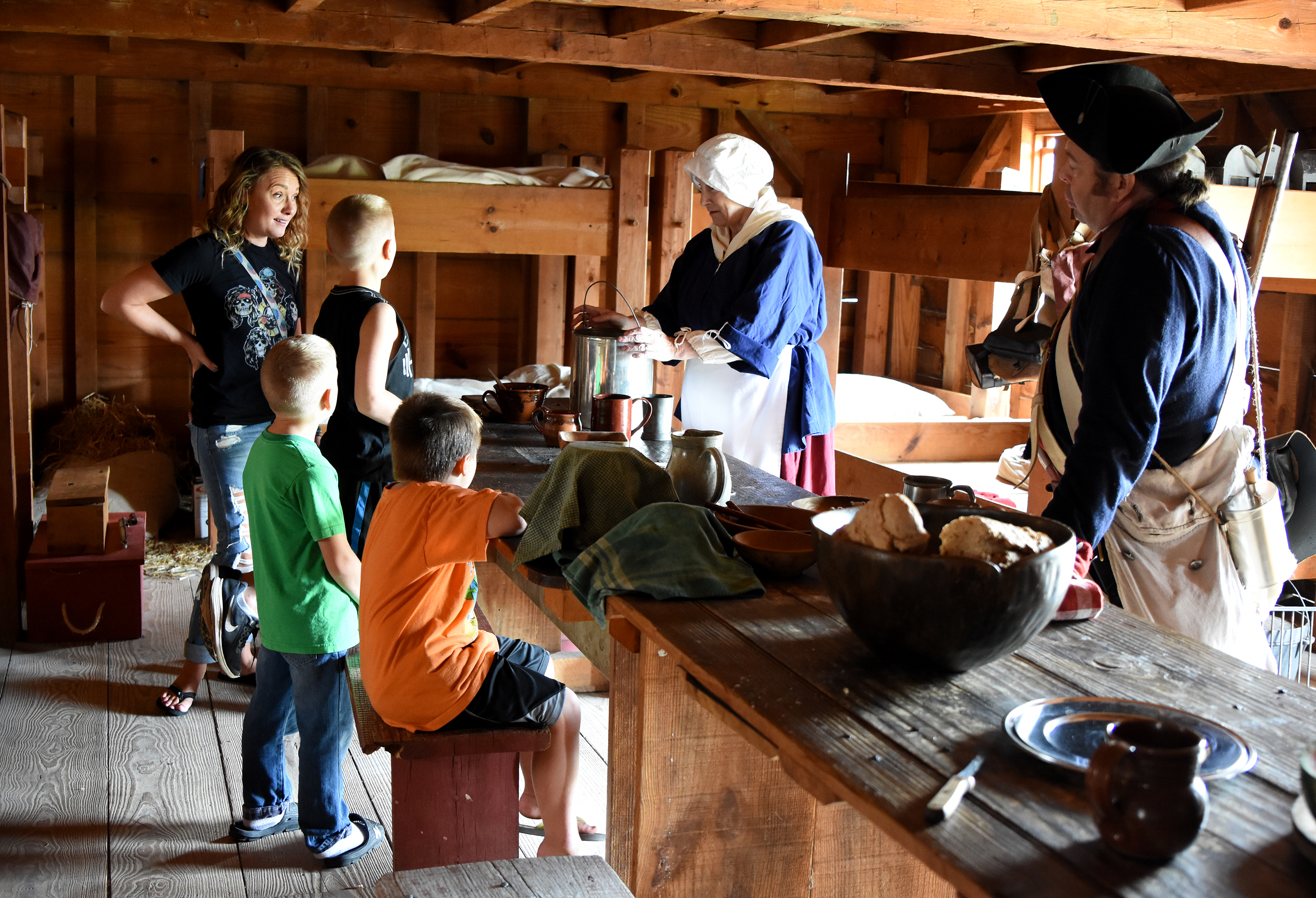 Children sits at a wooden table while speaking with a woman in 18th C clothing.