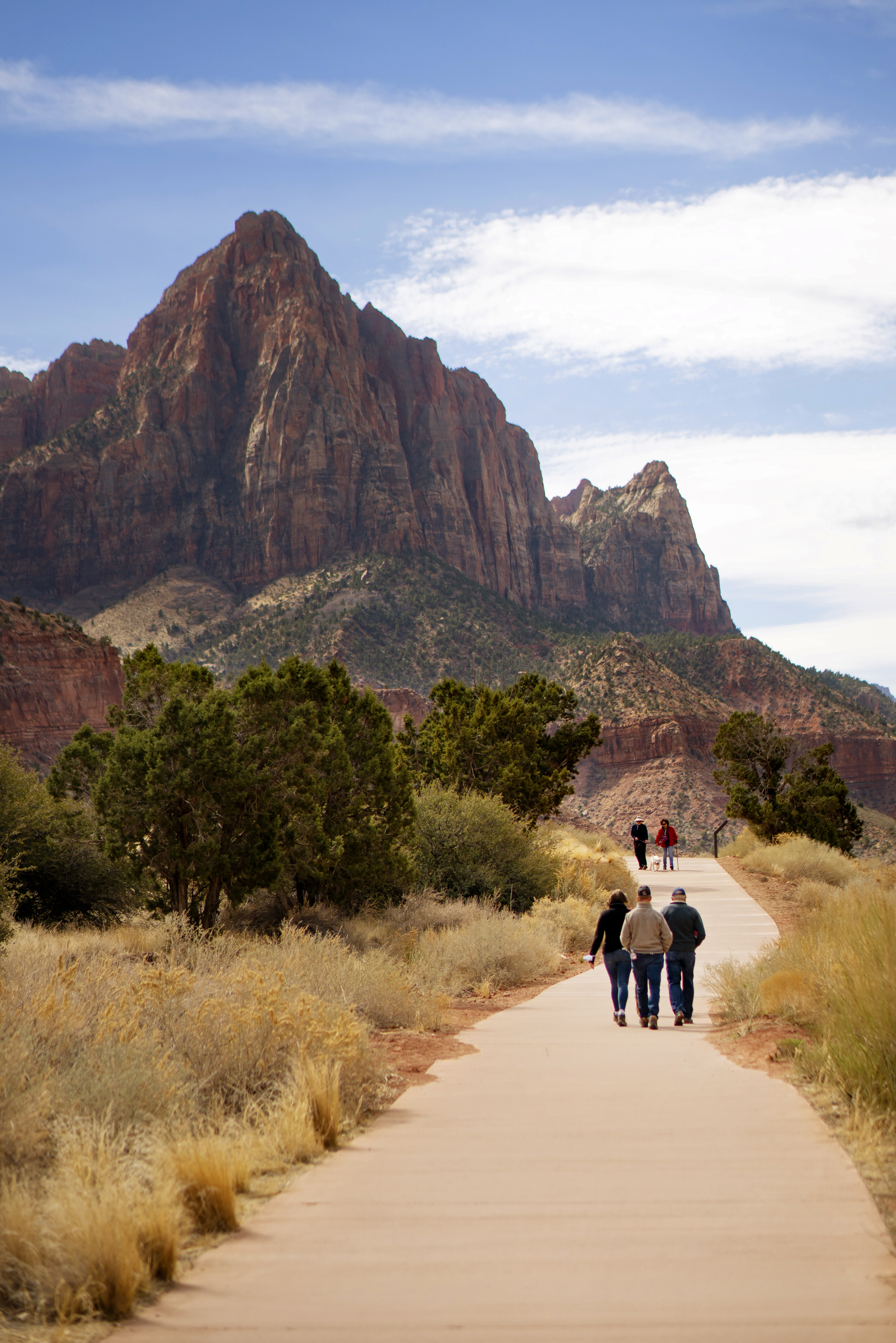 Three people walk on a paved path toward a triangular sandstone formation under a cloudy blue sky.