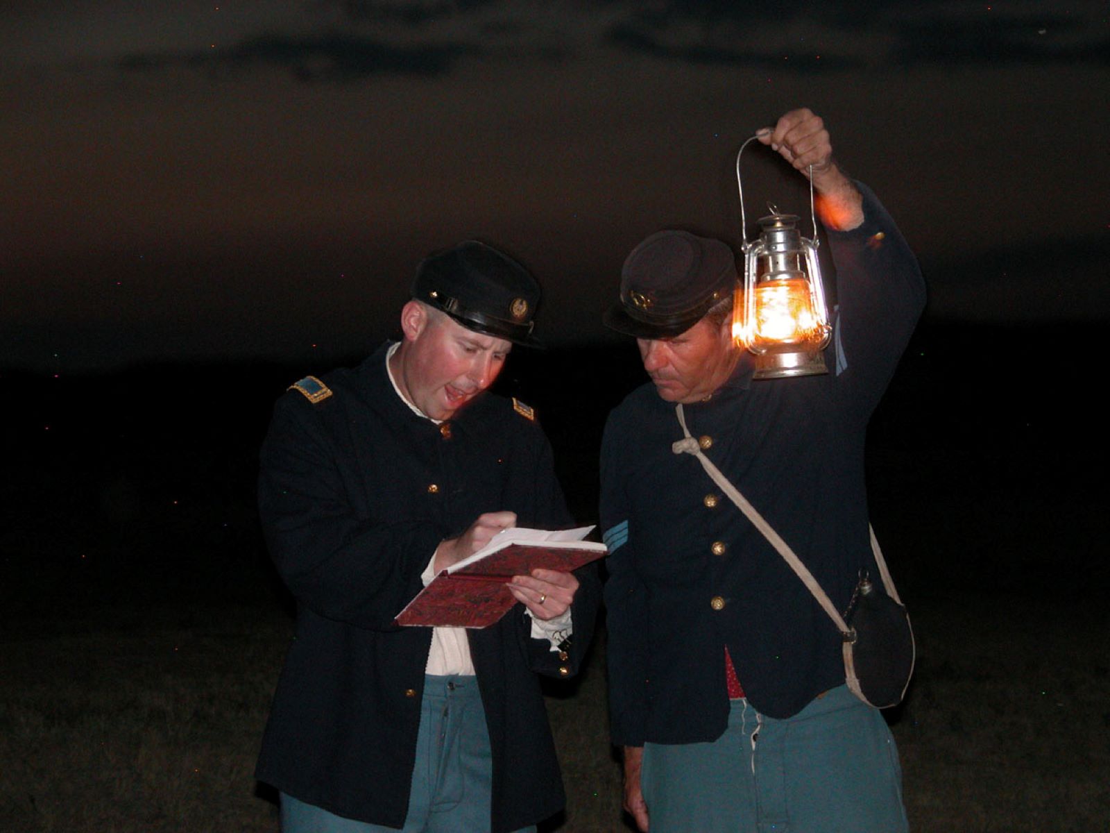Two soldiers dressed in blue uniforms stand next to each other reading a book by lamp light