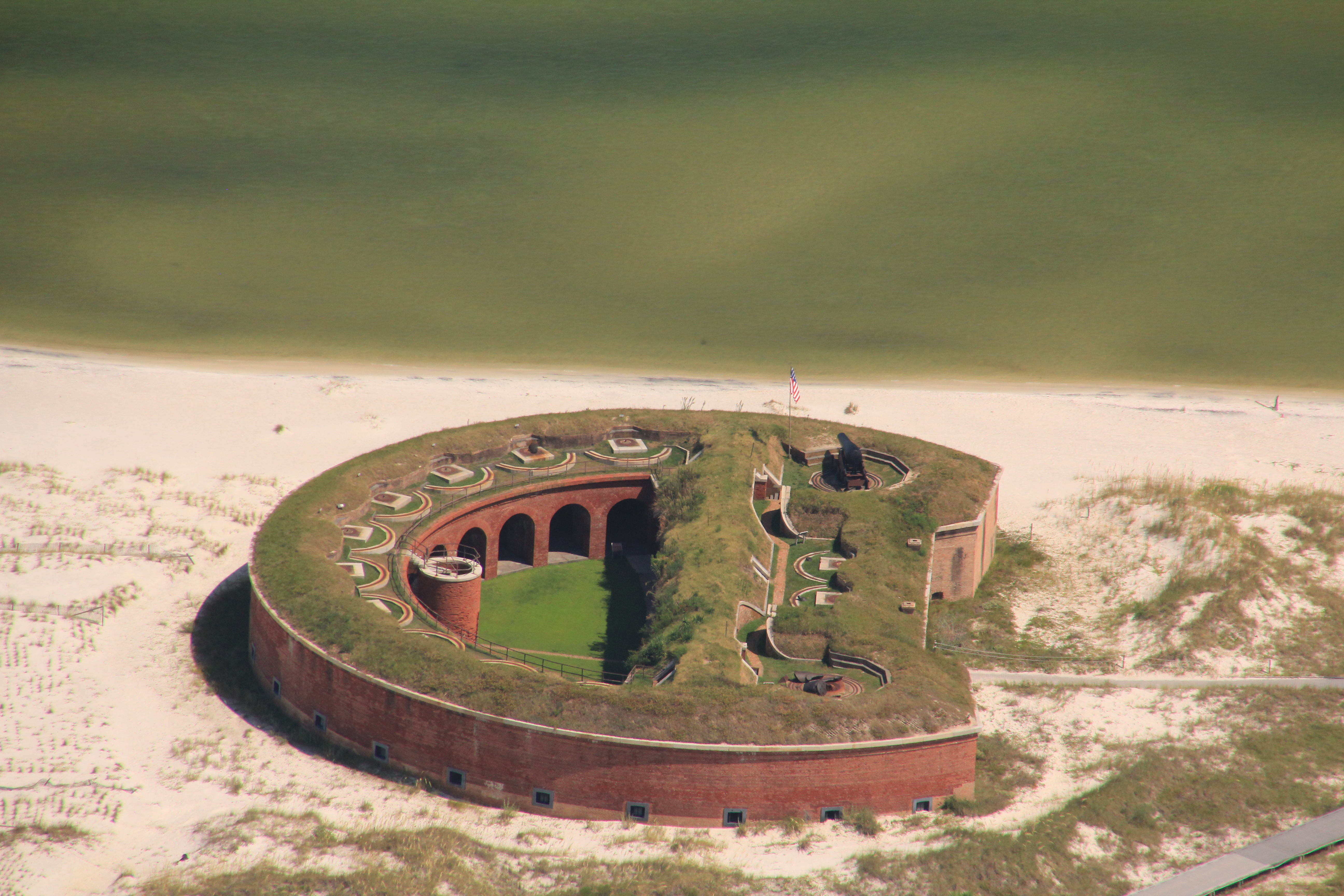 A brick fort stands against a blue sky with grasses and white sands at the entrance.