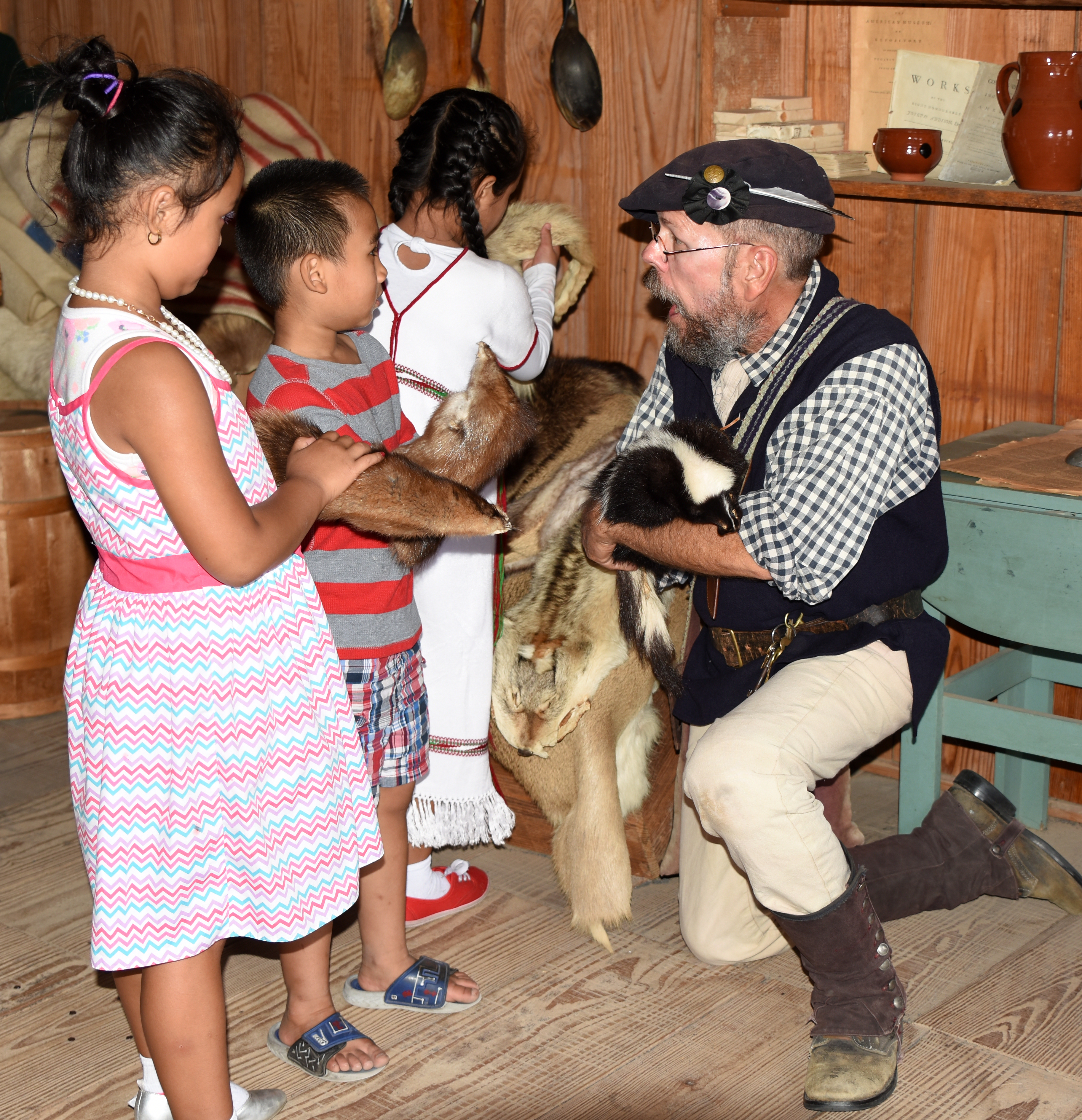 A Continental soldier kneels down to show 3 children fur pelts. He holds a skunk pelt.