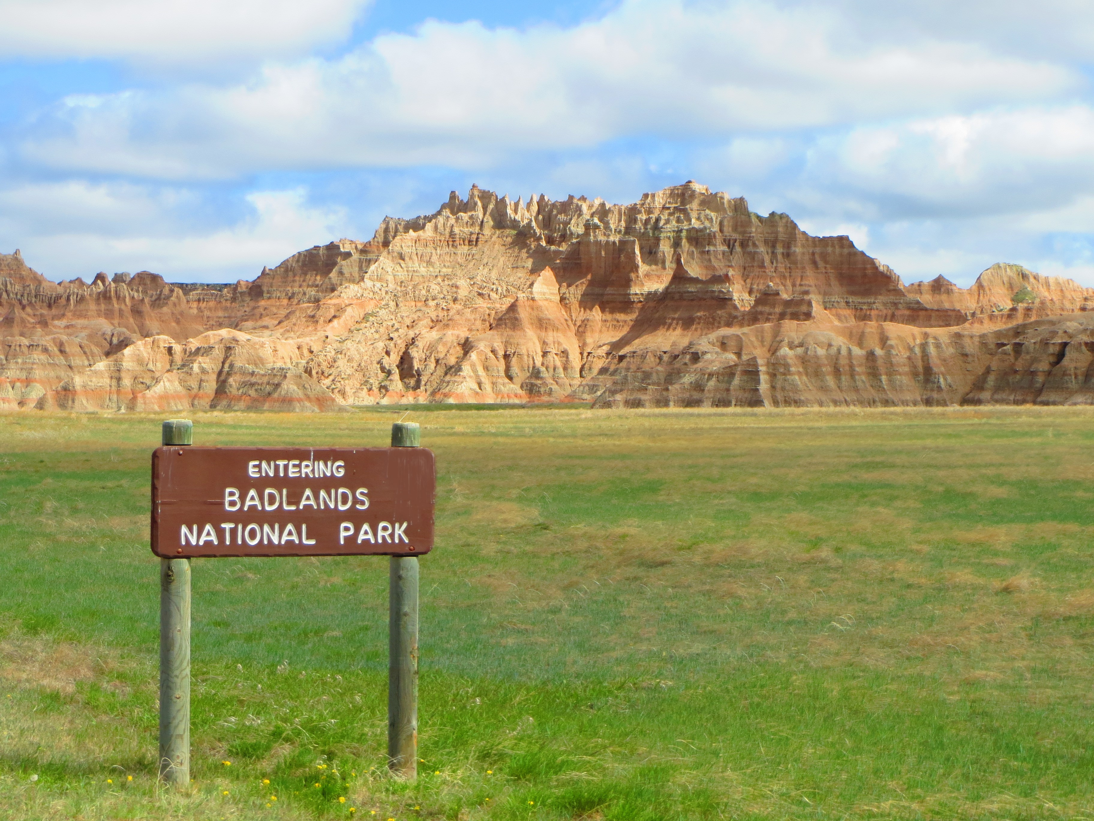 A brightly lit green field with badlands formations in horizon under blue sky.