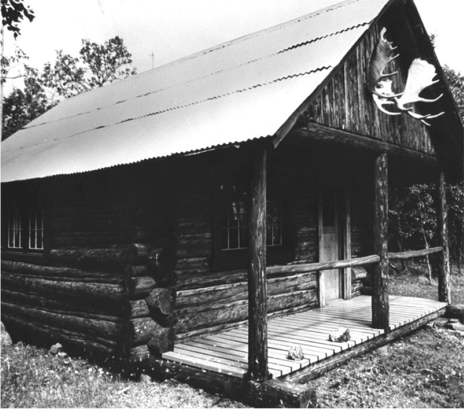 Black and white photo of log building with porch and roof and moose antlers hanging on top