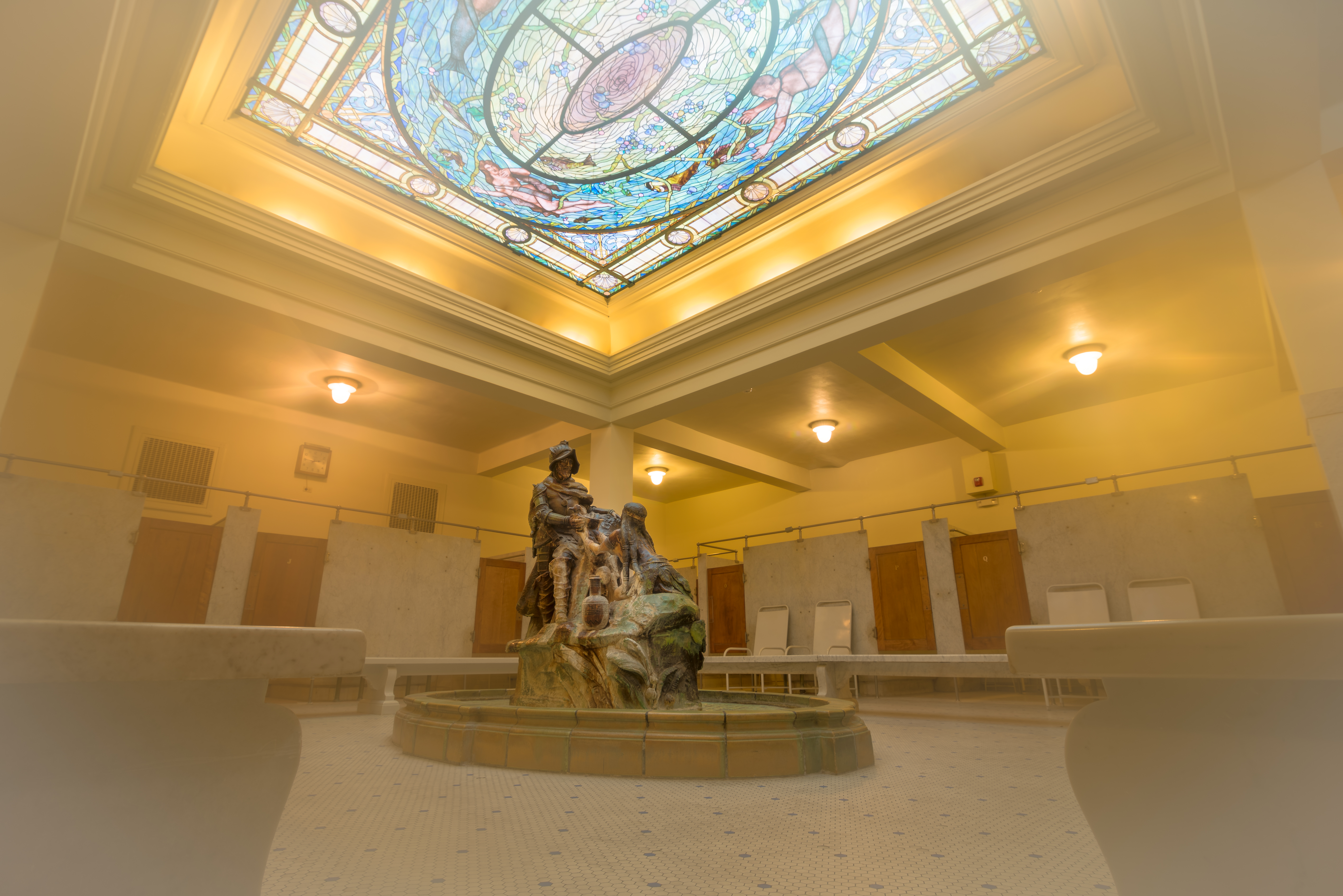 A statue with a man and a woman lies in the center of a room with marble furnishings and skylight.