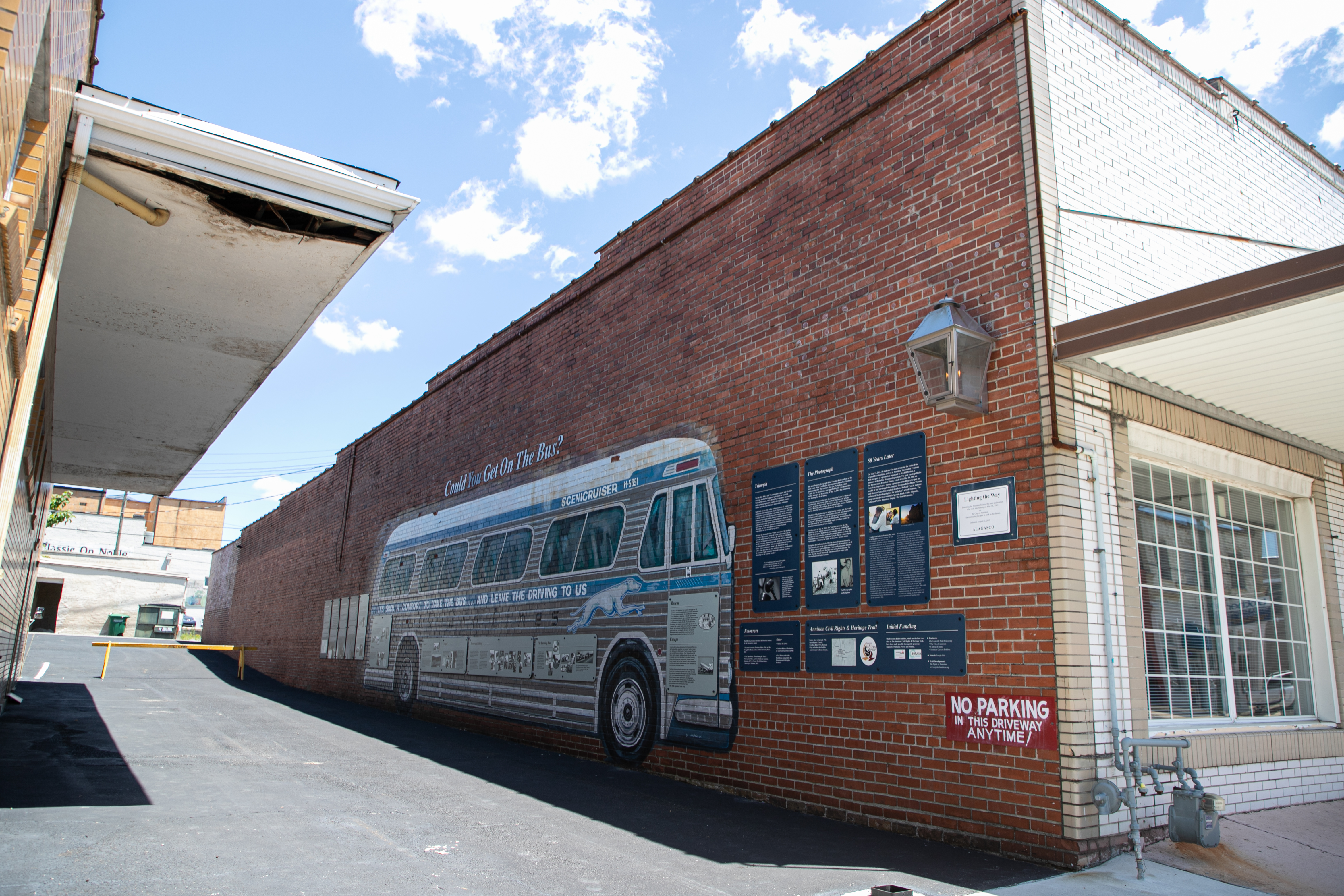 a red-brick wall facing an empty alleyway featuring a large silver painting of a bus and panels.