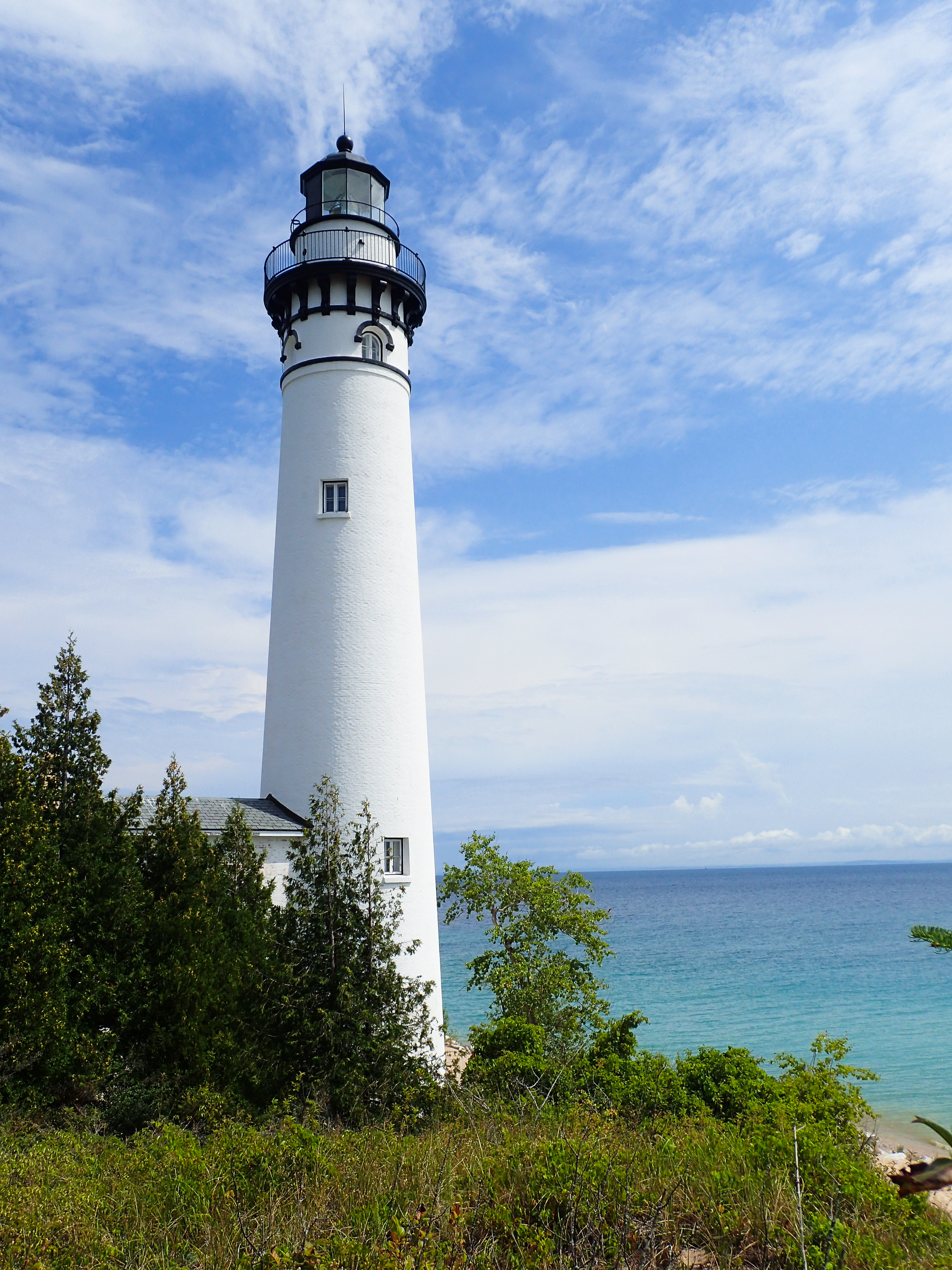 A white brick lighthouse sits on the edge of a large lake with blue skies beyond and shrubs