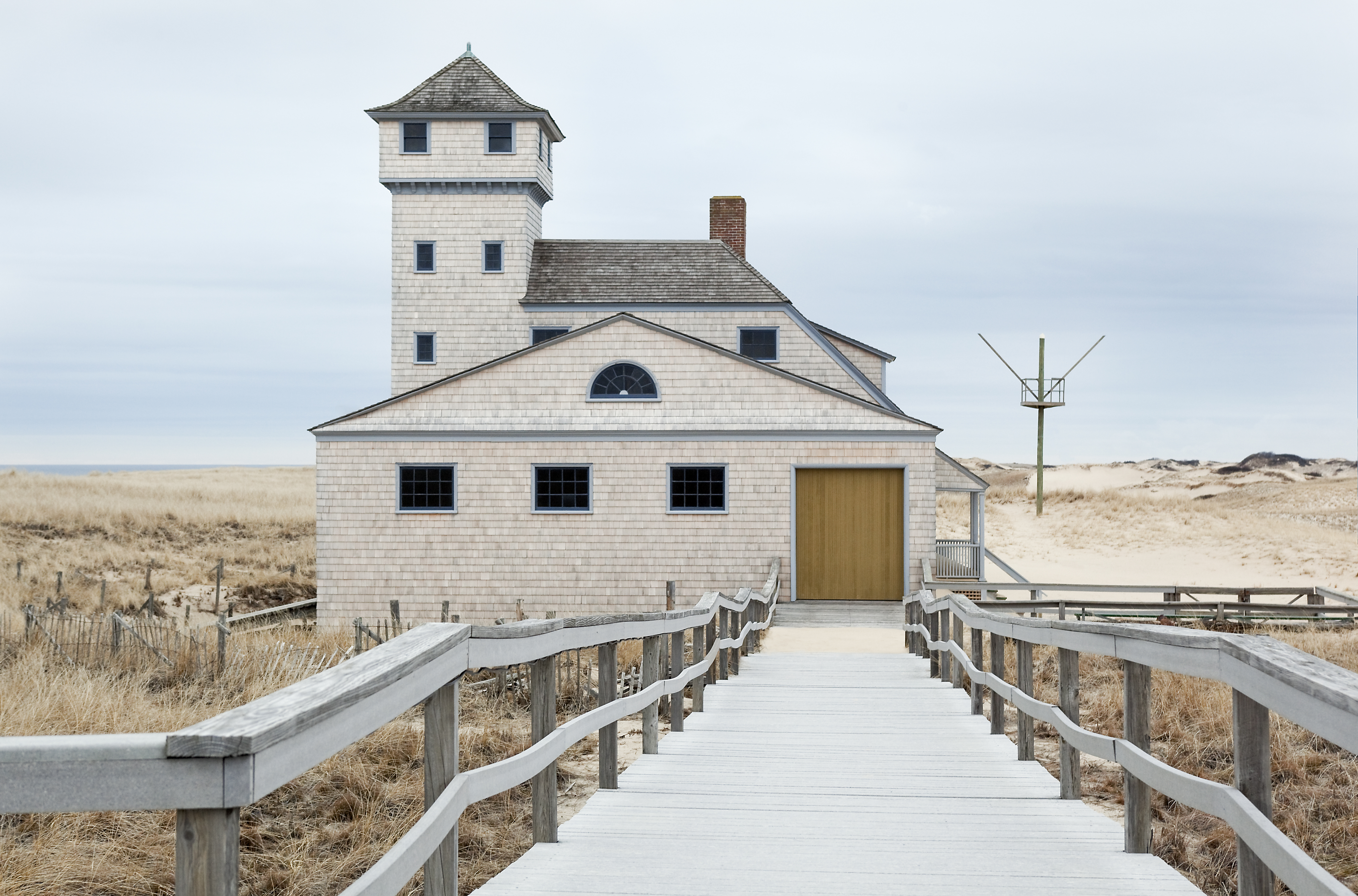 Ramp leads down to garage of Old Harbor Life-Saving Station. House against dunes and gray sky.