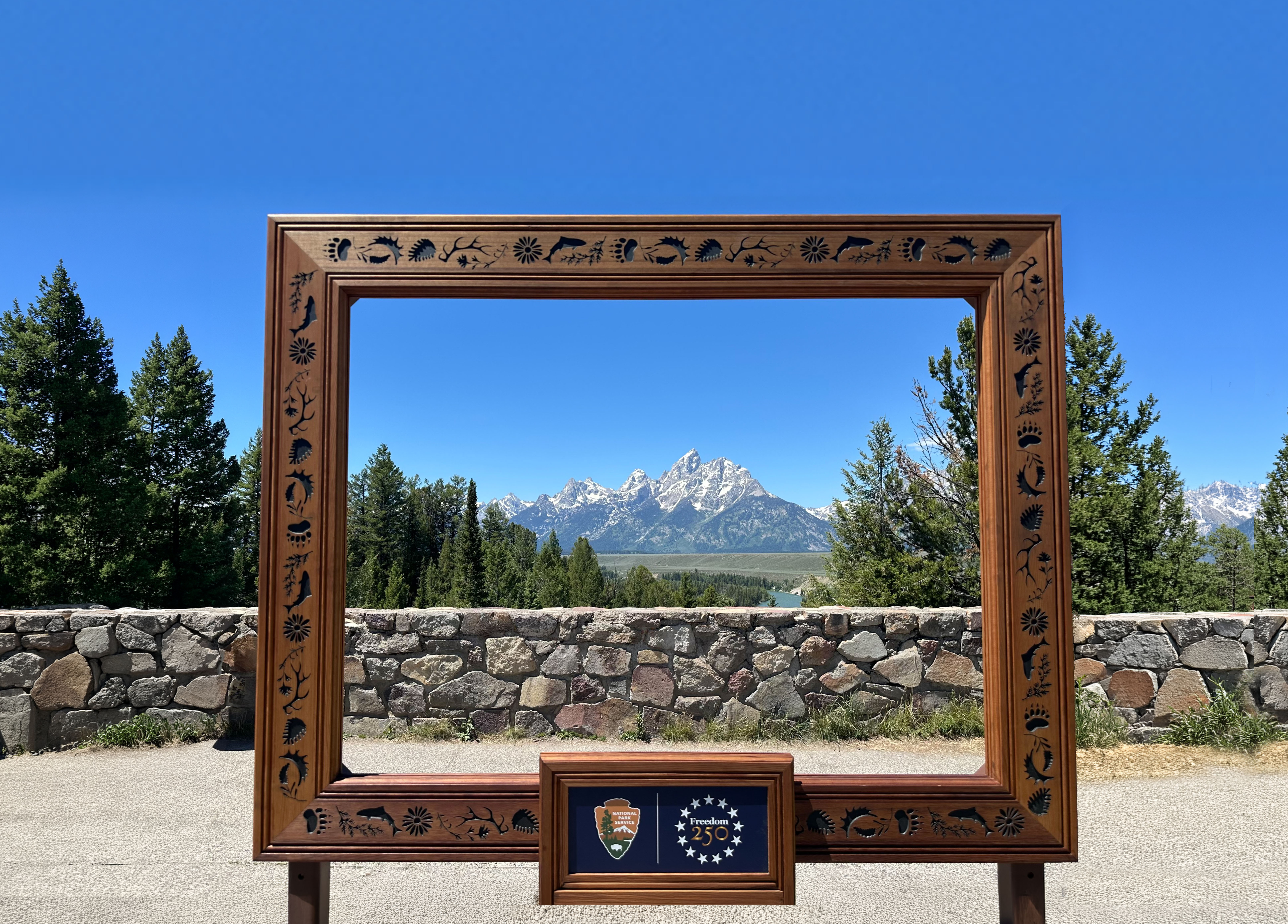 A wooden picture frame with a view of the teton mountains, blue sky, and conifer trees.