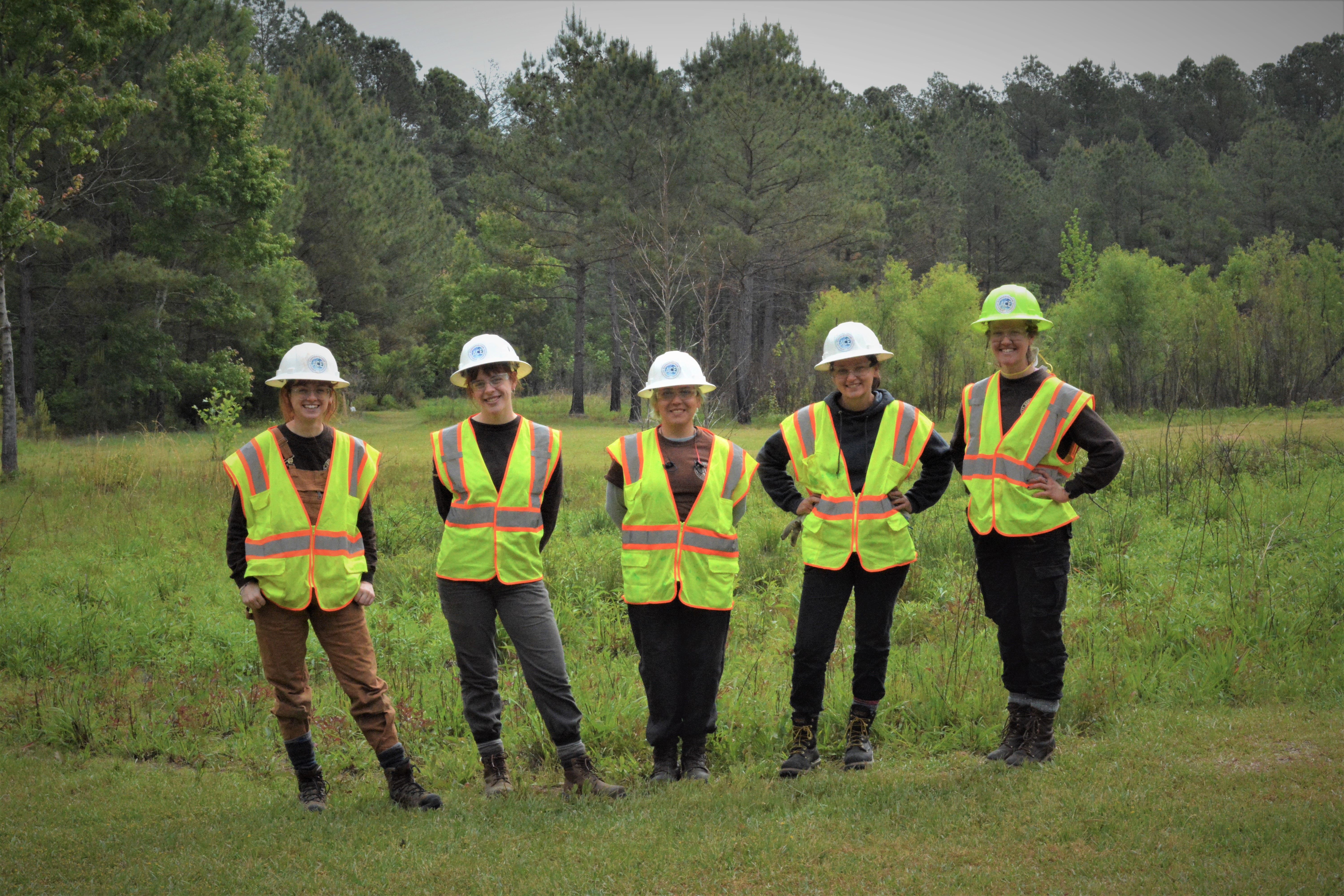 five people standing in line outside with personal protective equipment on; trees in background