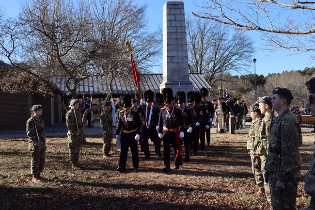 Men dressed in 18th century military uniforms with tall dark hats are marching in a line surrounded by people in camoflage holding swords high in honor to them.