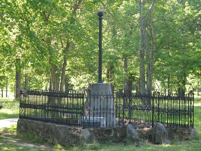 Smooth multi-sided concrete base topped with flagpole and cannon ball are surrounded by a black iron fence. Green-leaved trees are around it.