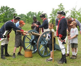 a group of men in military dress show a group of children an artillery drill in a green field with green trees in the background.
