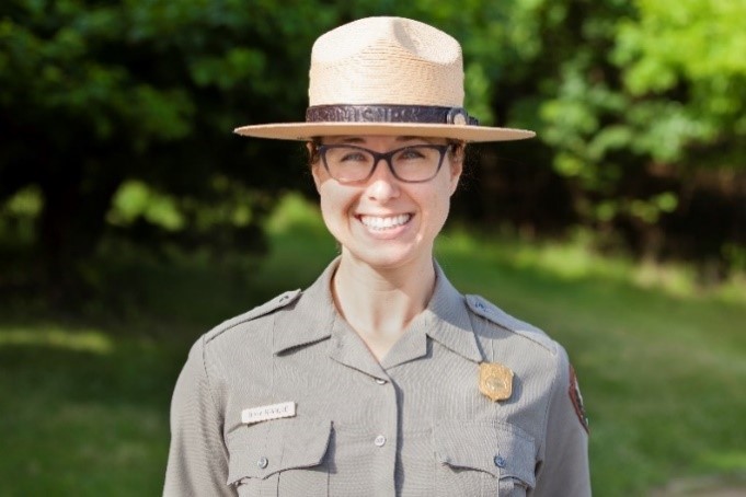 Diana Bramble, superintendent
Cowpens National Battlefield and Kings Mountain National Military Park Woman, wearing black-framed eyeglasses and a broad-brimmed hat, stands smiling in front of brilliant green foliage.