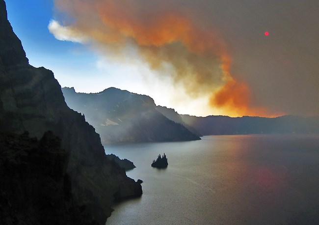 The eastern shore of Crater Lake and Phantom Ship are in the sunlight while an ominous cloud of smoke hovers above most of the lake. The smoke cloud edge is orange from the morning light.