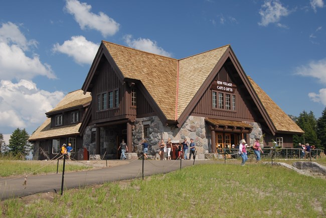 Stone steps lead to and people mingle around the historic two-story Rim Village Cafe & Gifts structure which has a four-pitched wood shingled roof, native stone first floor and brown painted wood slats on second story.