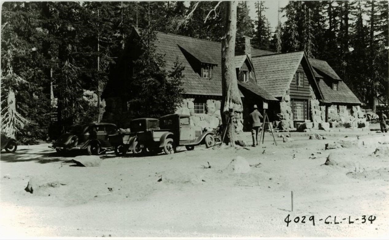 a 1934 black and white photo of a rustic style 2-story building constructed with native stone, wood shingles, and four dormer windows. Two men survey the front landscape. cars are parked on the side of the building