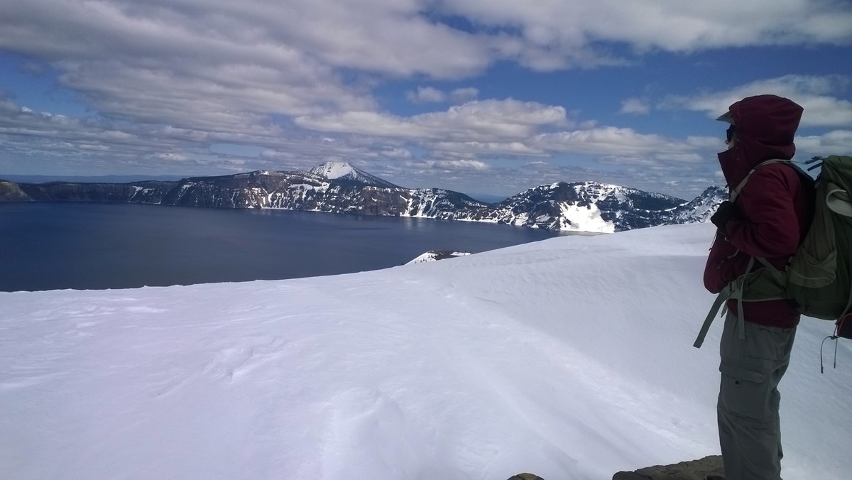 A person in a red winter coat with a backpack stands on a boulder looking across fresh snow on the rim surrounding  Crater Lake