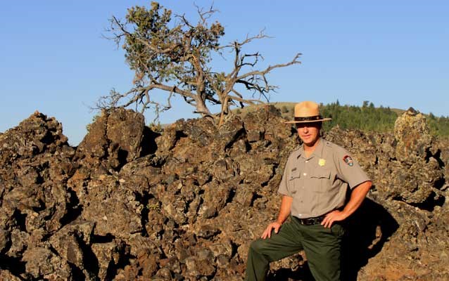 a ranger standing in front of a pile of black lava rocks with trees in the distance