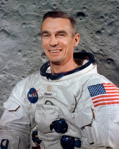portrait of a man in a white spacesuit with the American flag and NASA emblem in front of a photo of the moon's surface