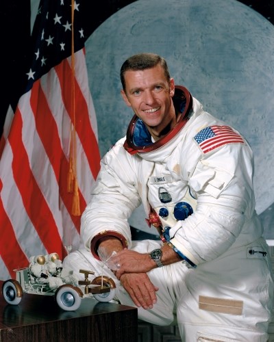 portrait of a man wearing a white spacesuit with several patches on it next to a model lunar rover