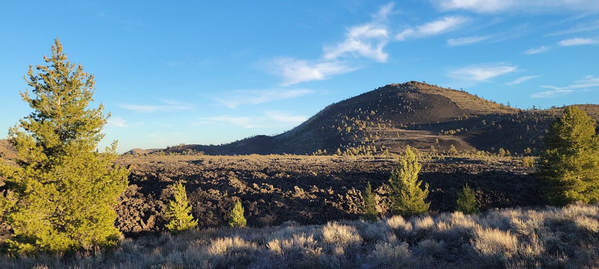 a large cinder cone in front of a dark lava field with trees and sagebrush