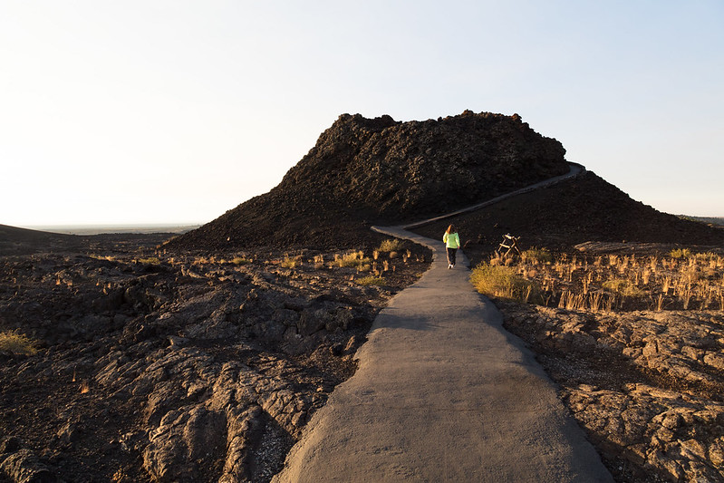 A black spatter cone rises from a lava landscape. A rugged black concrete trail leads to the base of the cone and slowly wraps around the cone's surface.