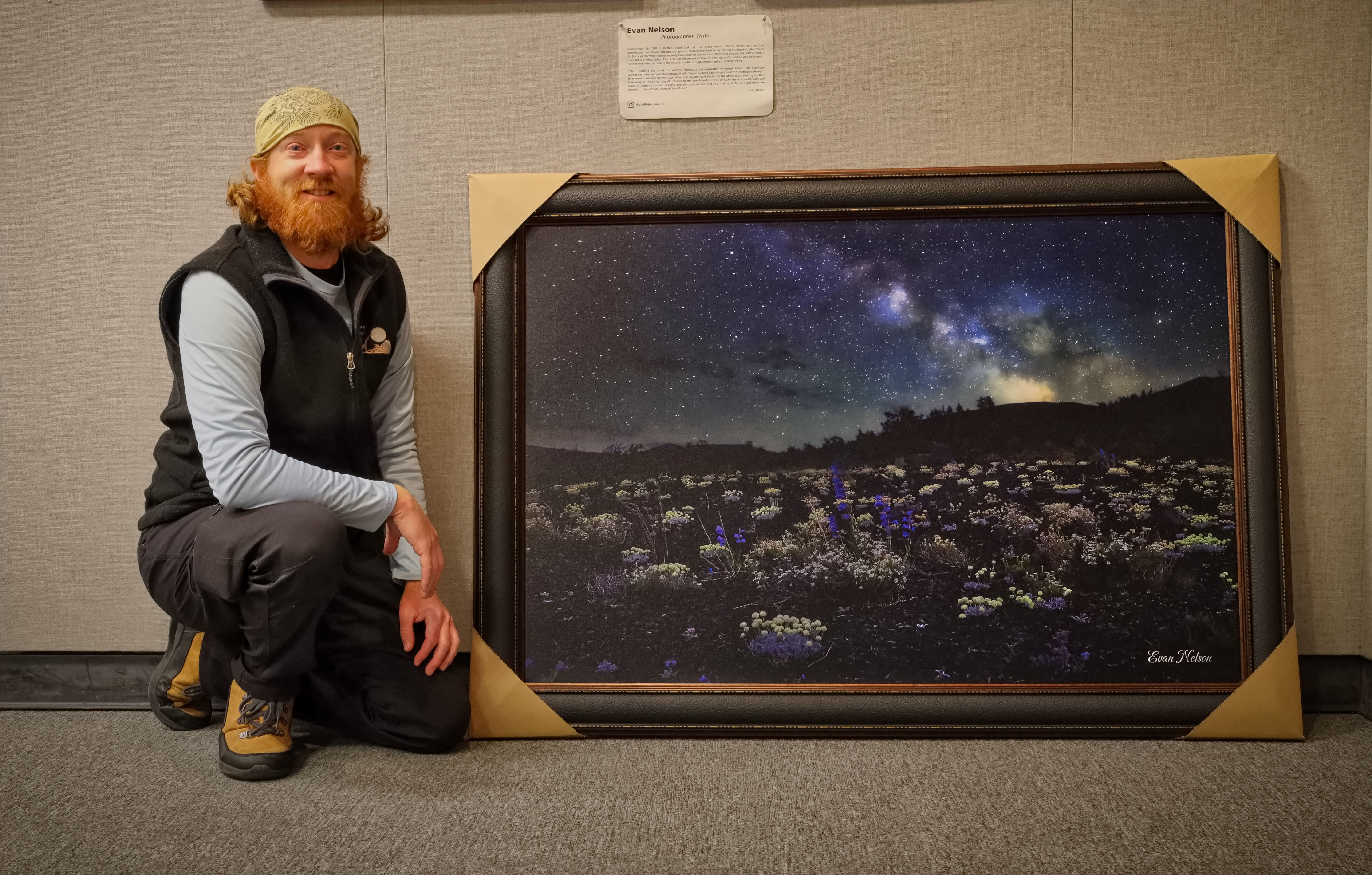 Man kneels next to a framed photograph of a a volcanic landscape with wildflowers illuminated byt the stars and Milky Way.