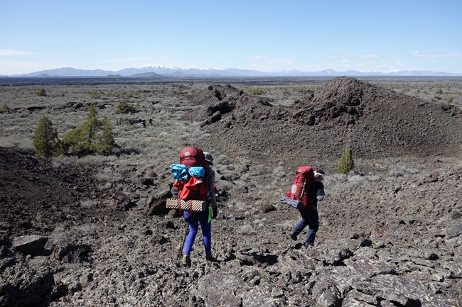 two hikers with backpacks hiking across a rocky landscape with few plants and mountains in the distance
