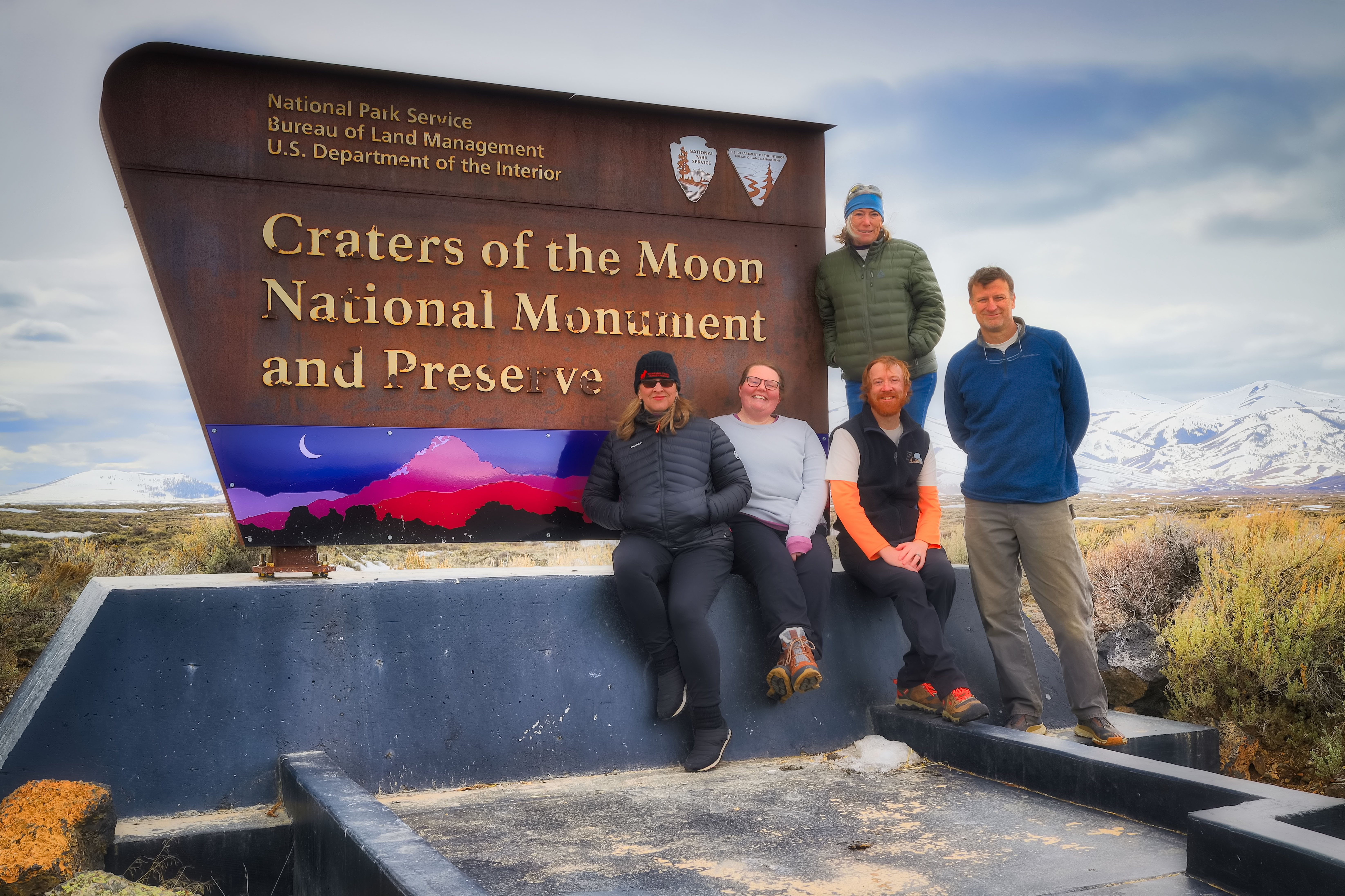 AiR Group Photo Group of people sit and stand in front of the entrance sign for Craters of the Moon National Monument